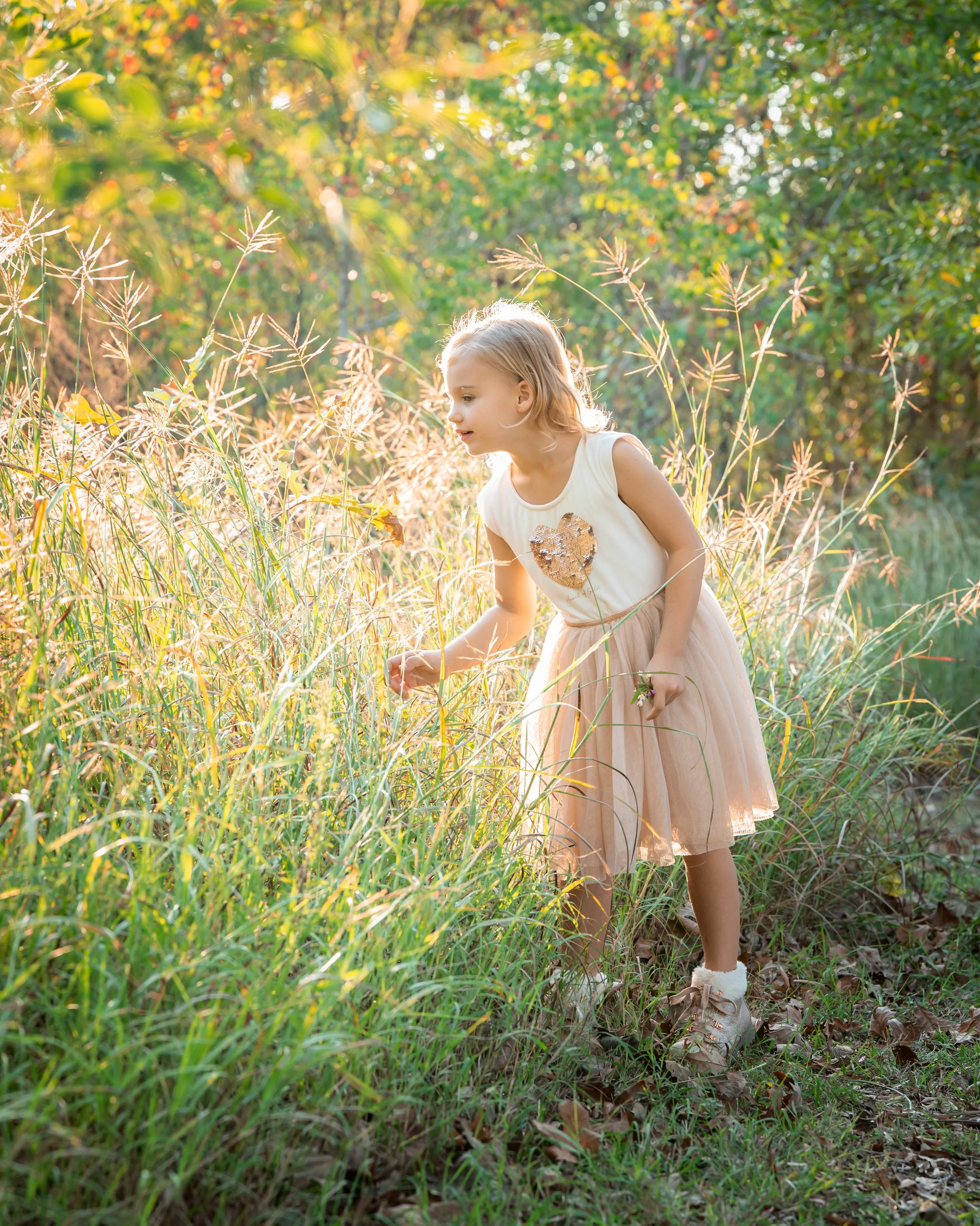 A young girl with blonde hair, wearing a sleeveless cream dress with a sequined heart, crouches in a field of tall grass and wild plants during sunset, with green trees in the background and sunlight filtering through.