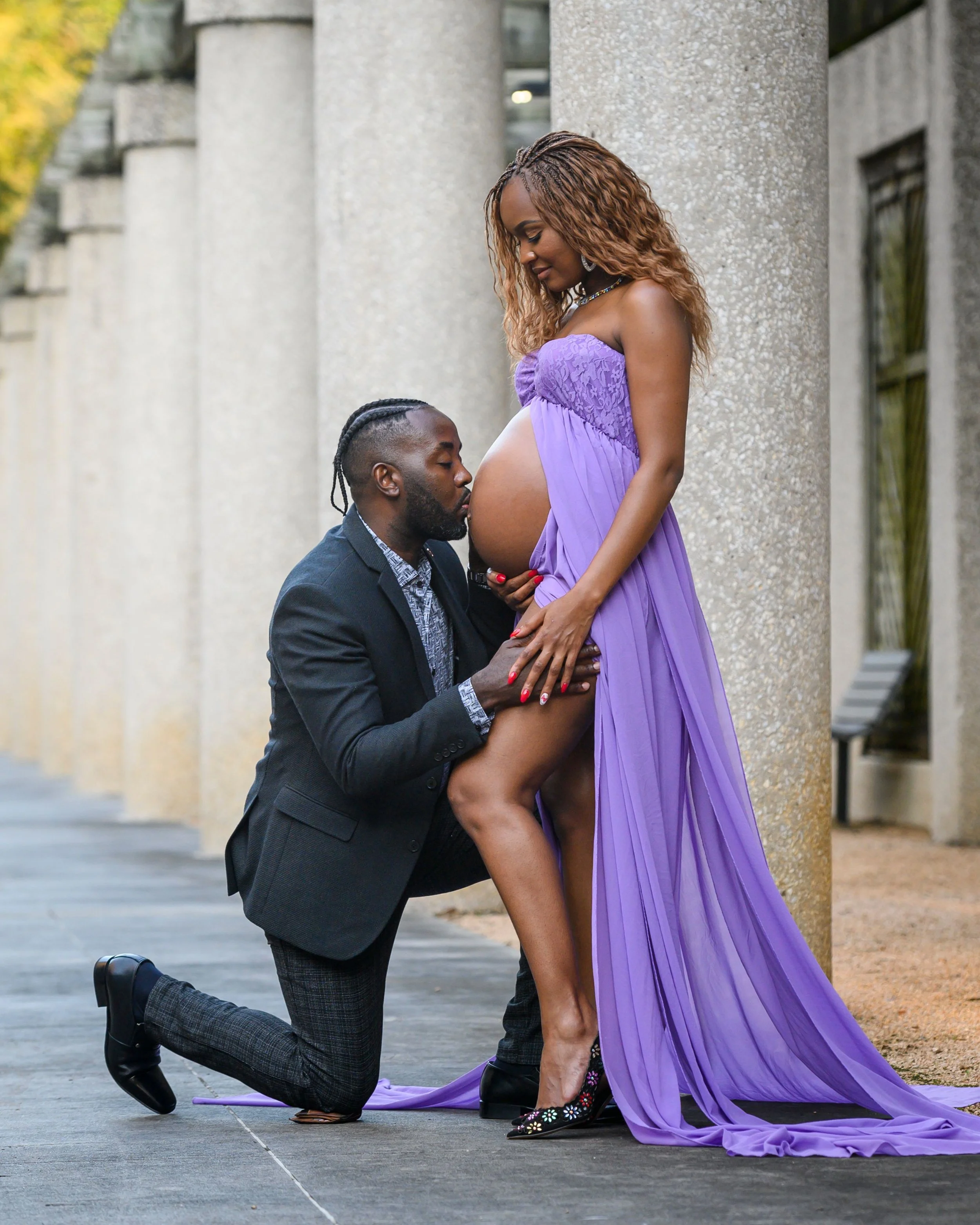 A man kneeling on one knee holding a pregnant woman by the waist, who is standing and resting her hands on her belly, in front of a row of large concrete pillars outdoors, with greenery visible in the background.