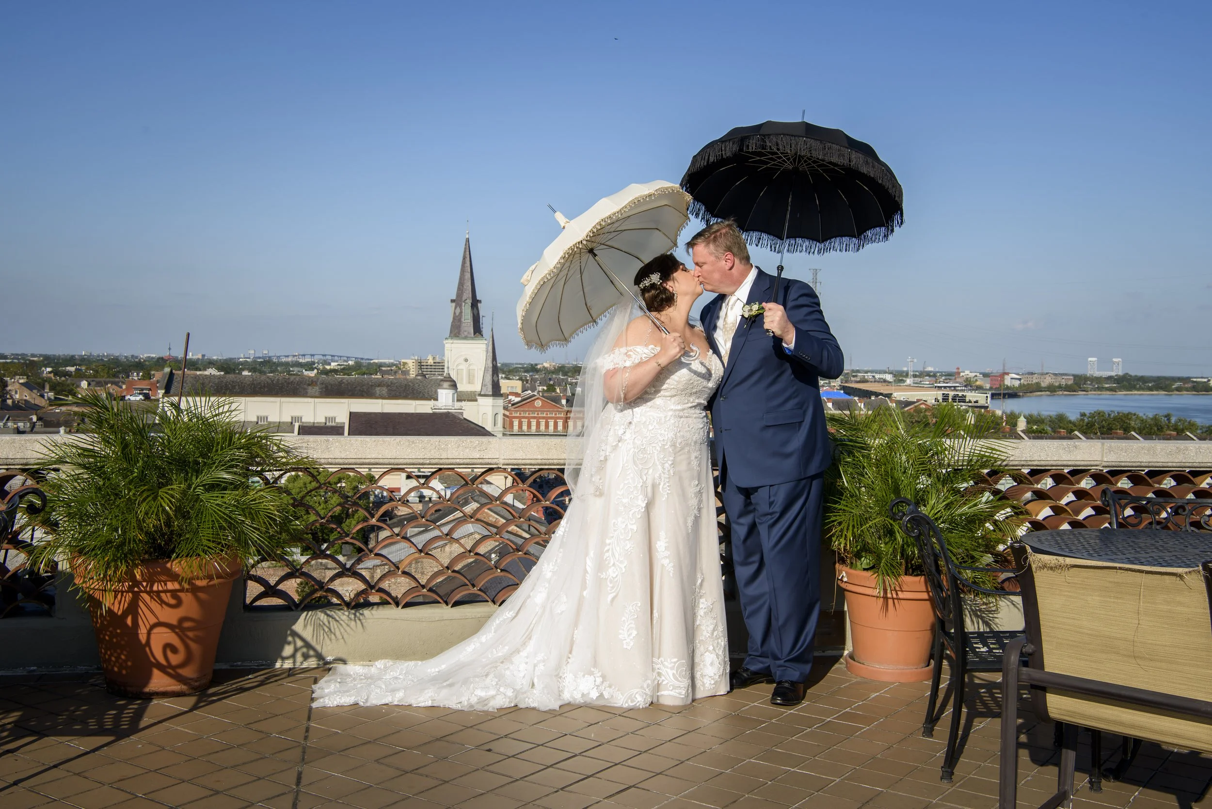 A bride and groom in wedding attire sharing a kiss under umbrellas on a rooftop with St. Louis Cathedral in New Orleans and a river in the background.