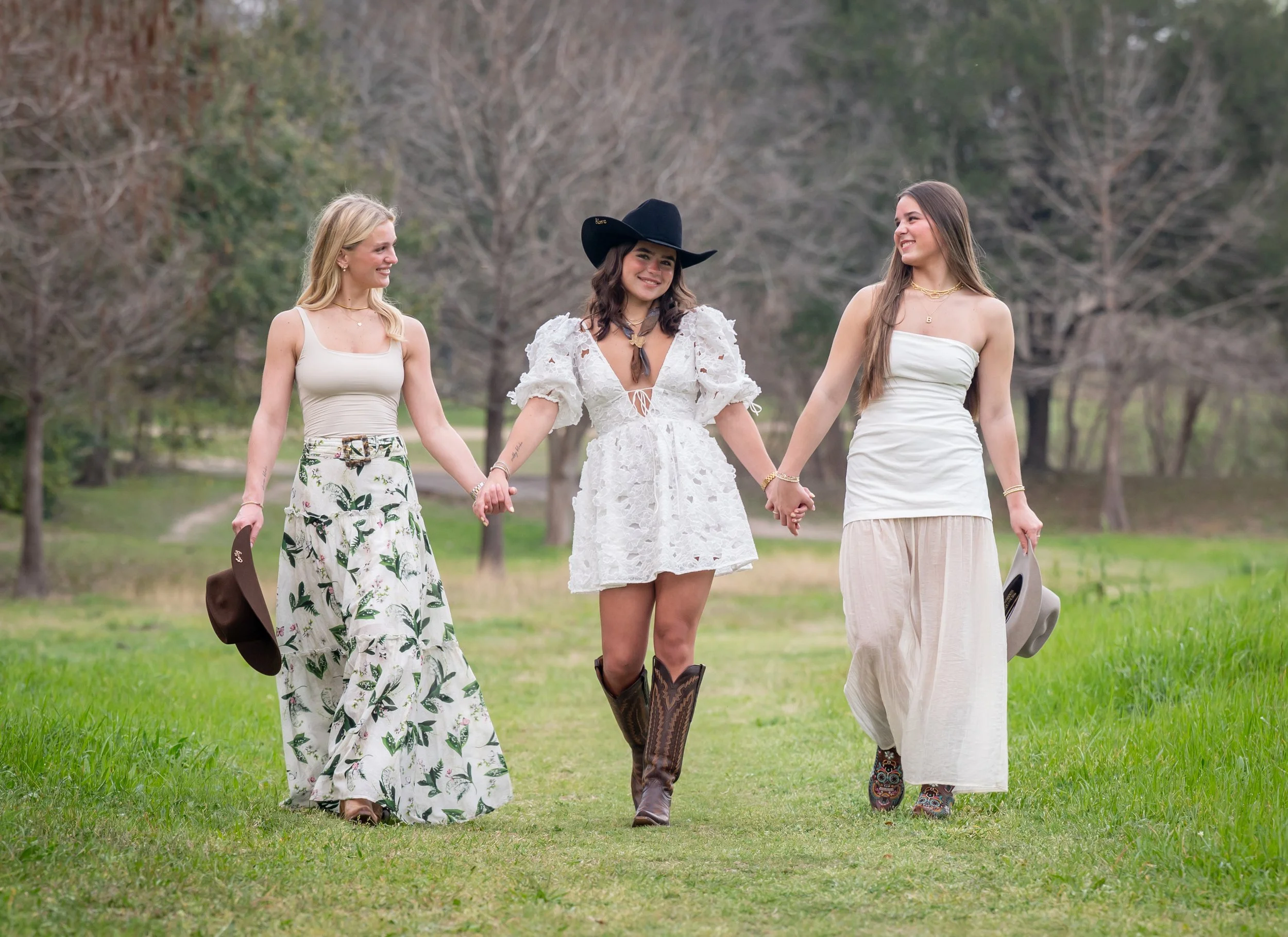 Three women holding hands and smiling while walking on a grassy outdoors area with trees in the background. The middle woman is dressed in a white dress and cowboy hat, and the other two women are wearing casual dresses.