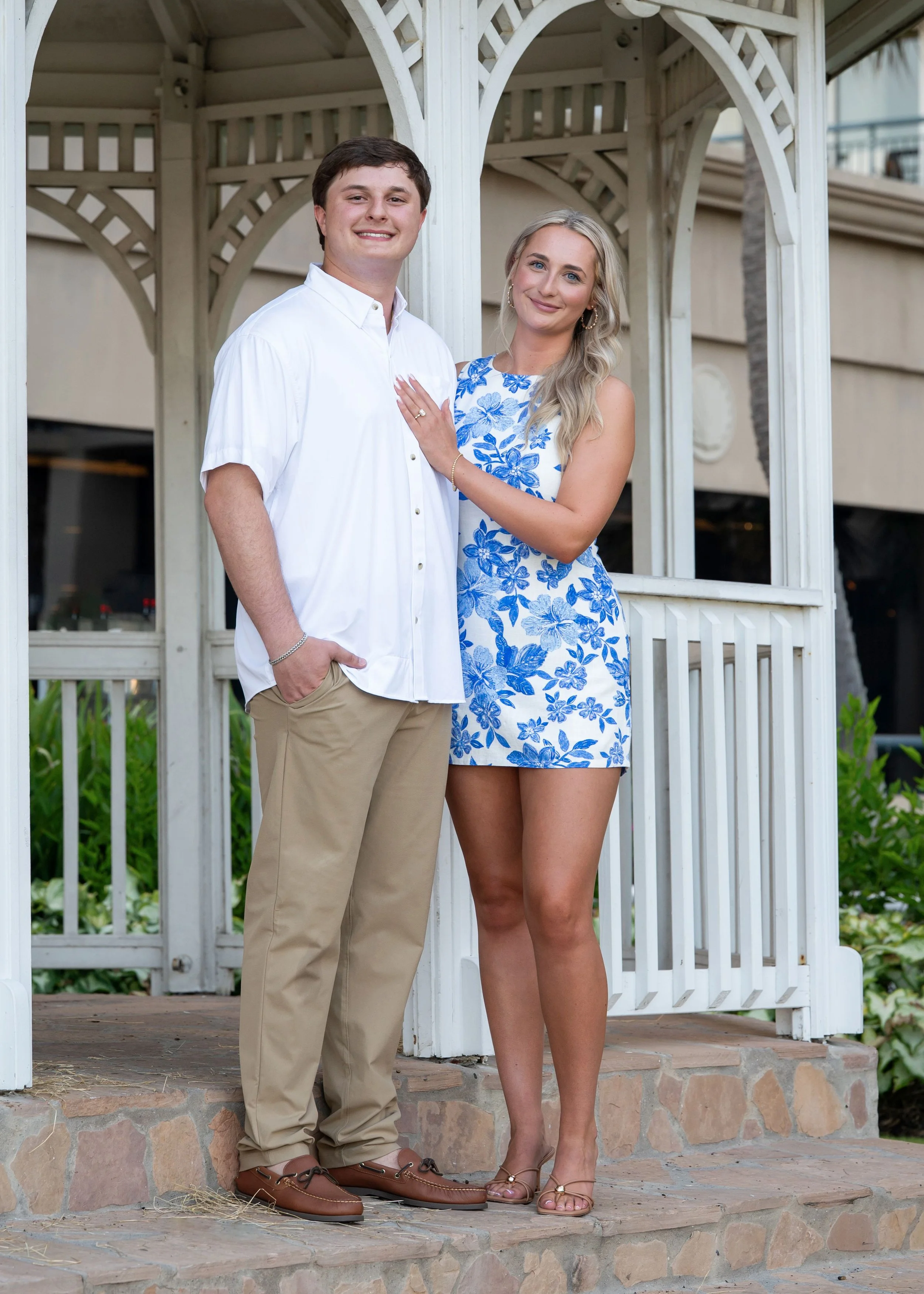 A young couple standing on steps in front of a white gazebo. The man is wearing a white button-up shirt and khaki pants, smiling with one hand in his pocket. The woman is wearing a blue and white floral dress, smiling with her hand on the man's chest
