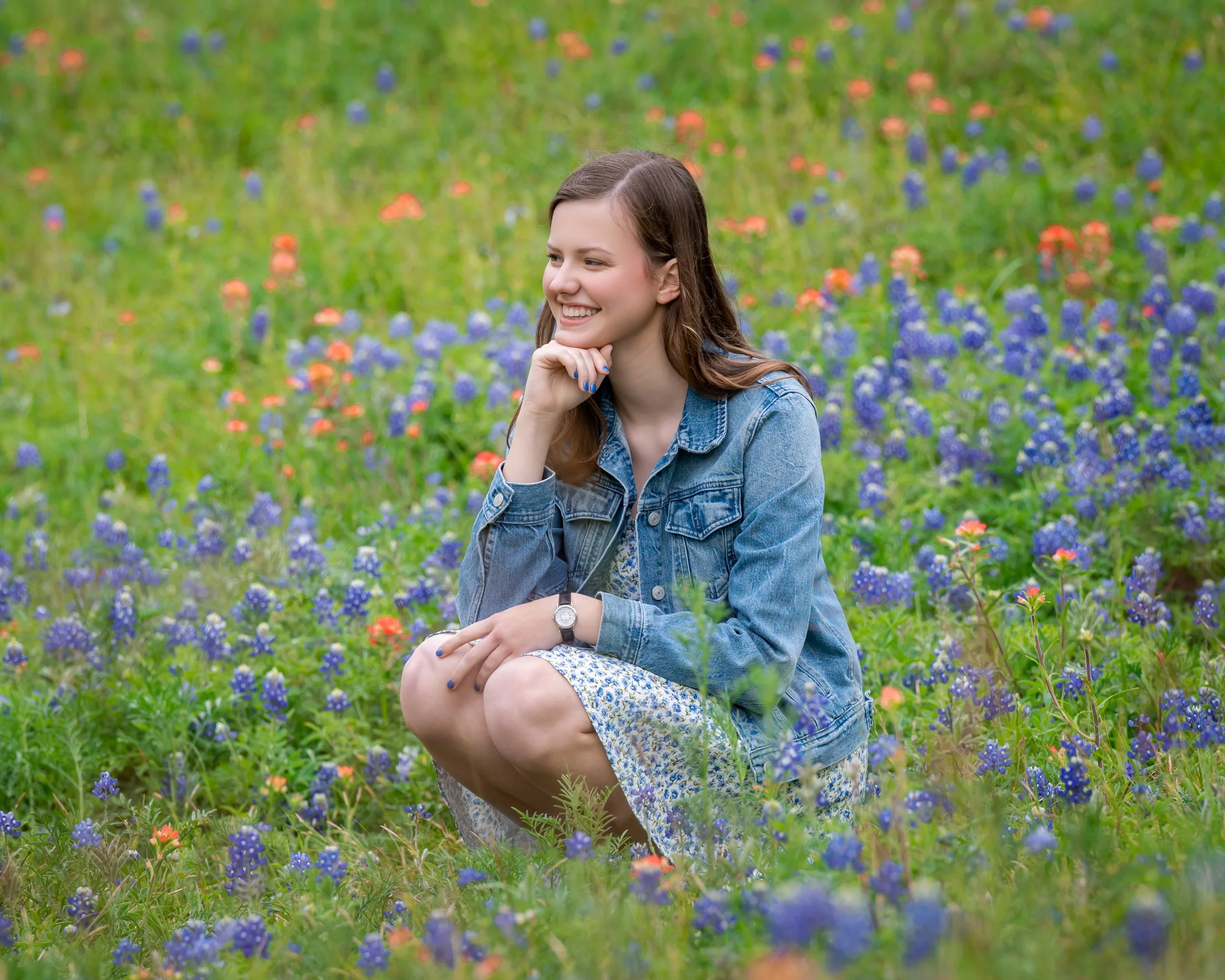 A young woman with brown hair, smiling, sitting among bluebonnets, wearing a denim jacket and floral dress, in a field of purple and orange flowers with green grass.