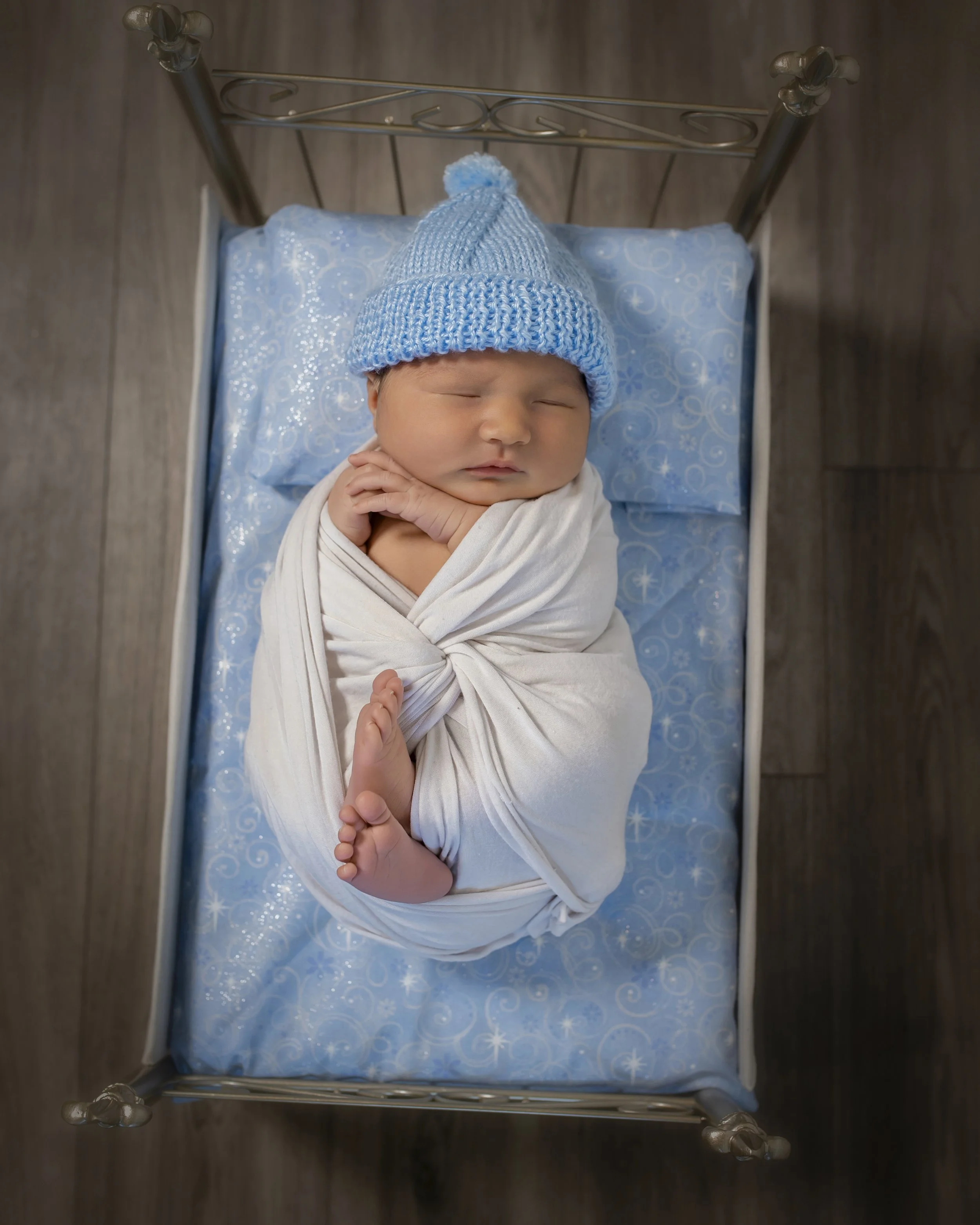 A sleeping newborn baby wrapped in a white blanket on a blue sheet with snowflake patterns, wearing a blue knit hat with a pom-pom, lying in a small crib.