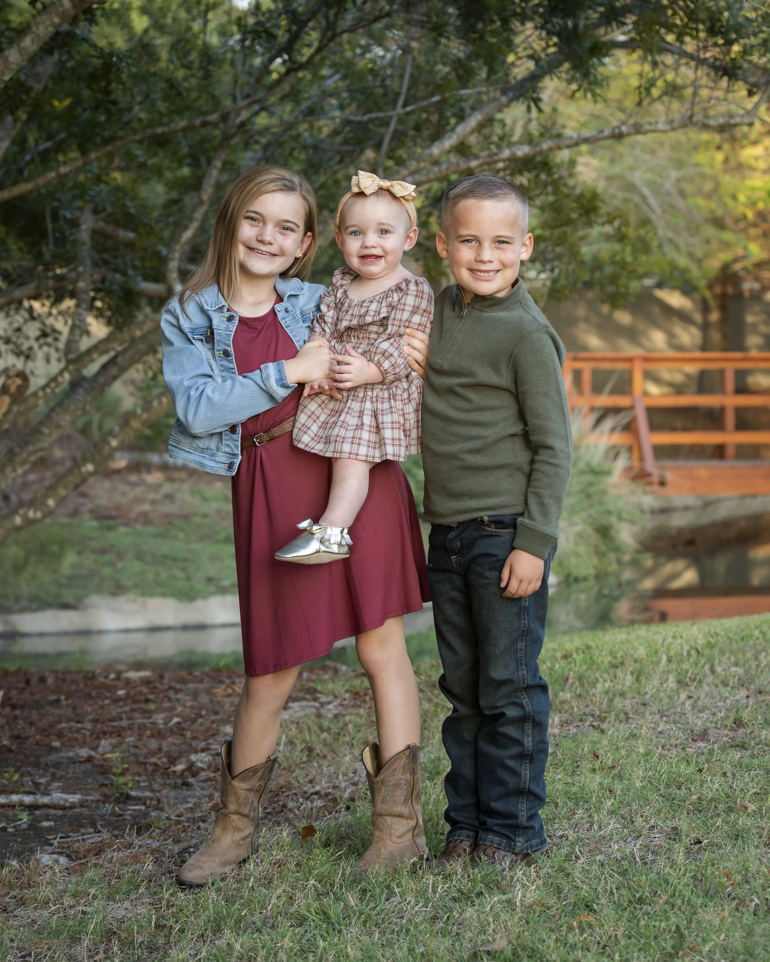 Three children standing outdoors in a park with trees and a wooden fence in the background. The youngest girl in the middle is being held by the girl on the left and the boy on the right. They are all smiling and dressed casually.
