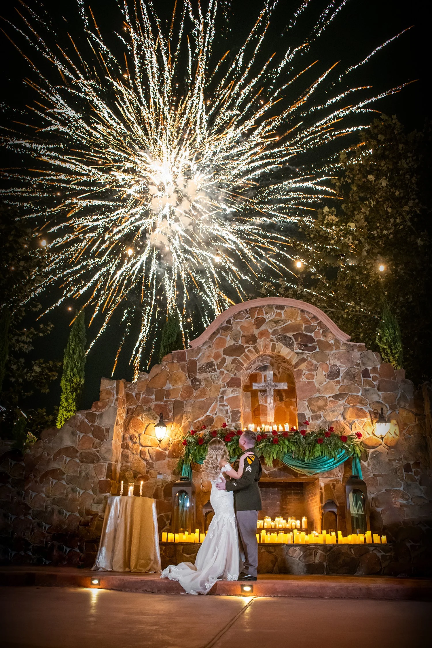 A bride and groom embrace during their wedding ceremony at an outdoor altar with a stone backdrop and burning candles, while fireworks illuminate the night sky above.