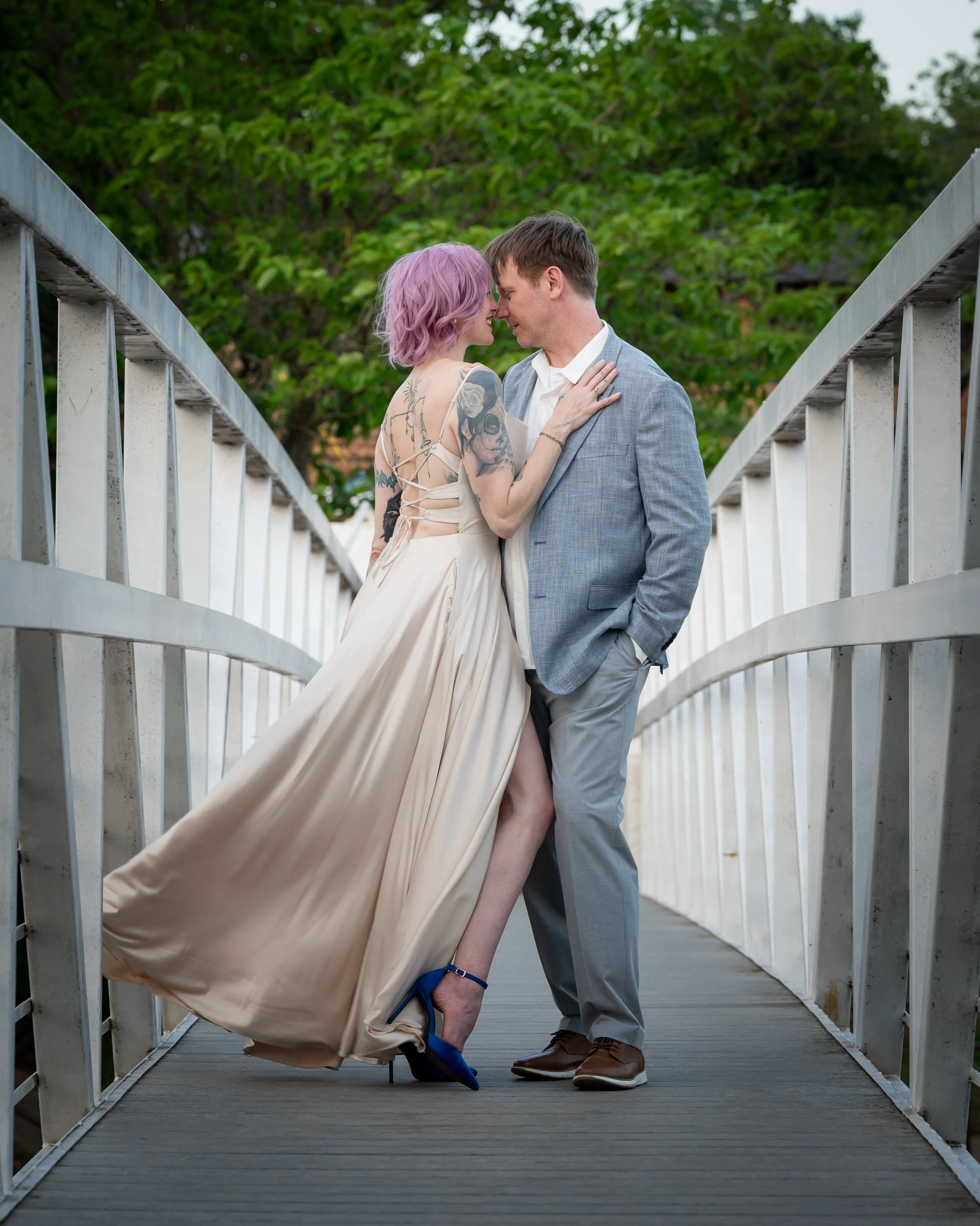 A couple on a bridge leaning in and touching foreheads, with the woman lifting her leg slightly and wearing a dress, shoes, and having tattoos, while the man wears a gray suit and brown shoes, surrounded by trees.