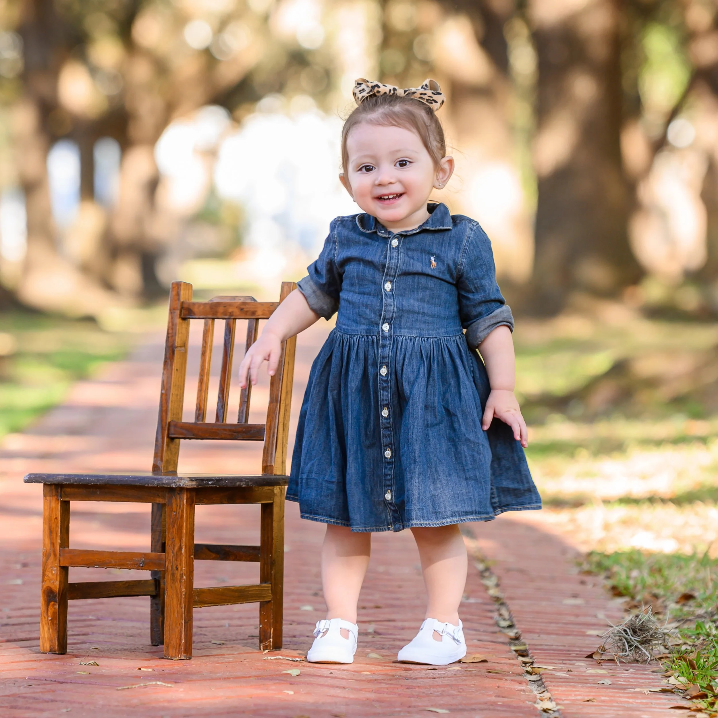 A young child with a leopard-print bow in her hair and a denim dress smiling and standing outdoors next to a wooden chair on a brick pathway in a park or garden.