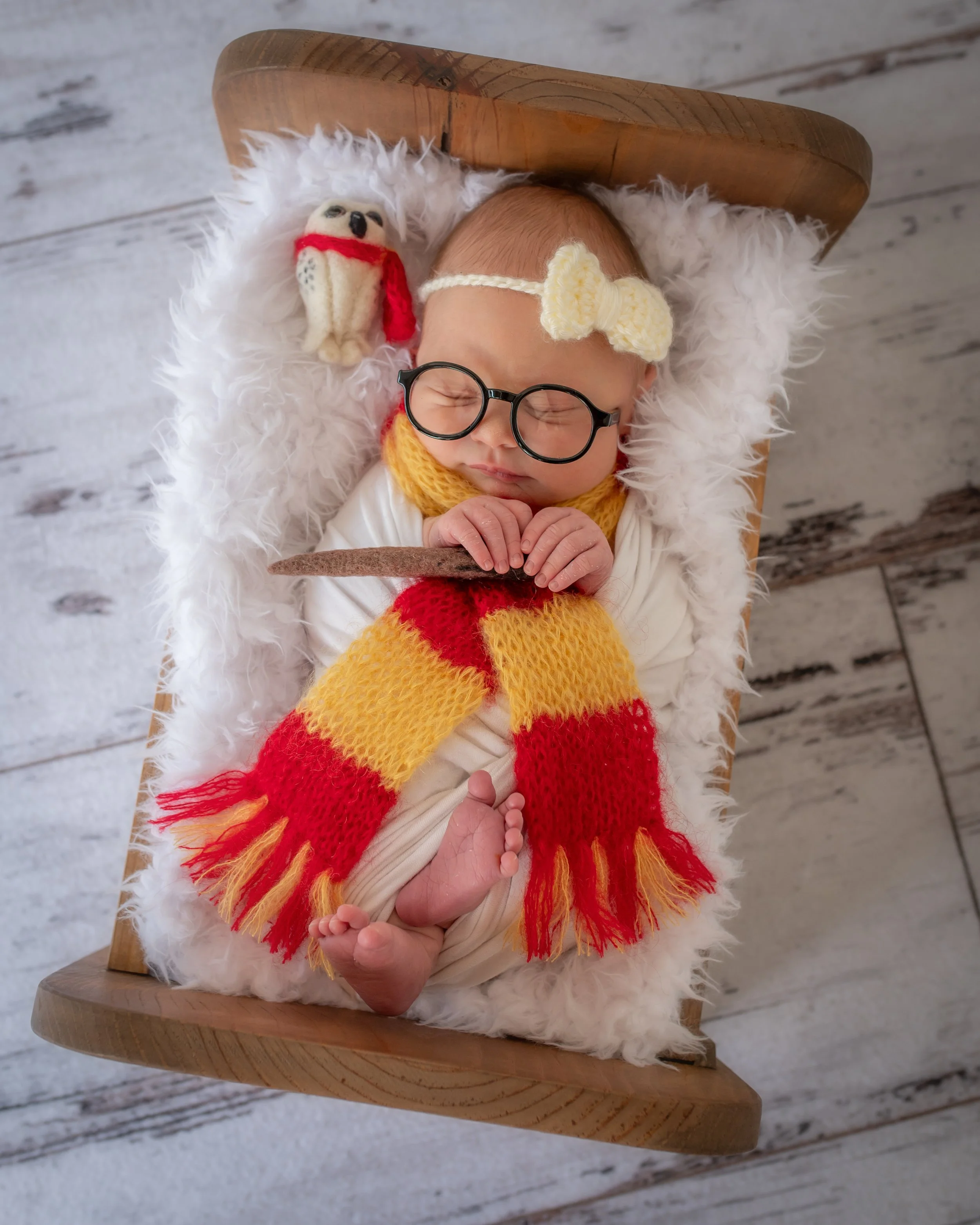 A newborn baby wearing glasses and a crocheted headband with a flower, sleeping on a fluffy white blanket in a wooden bed, with a small plush owl toy wrapped in a red scarf nearby. Harry Potter 