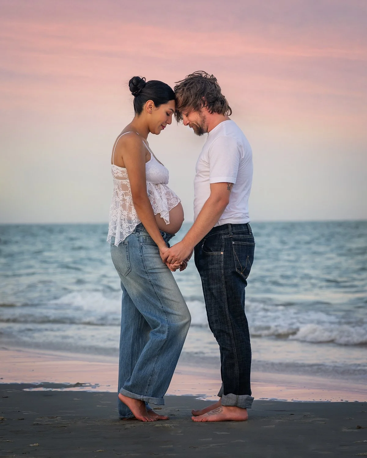 A pregnant woman and a man standing on Galveston beach, holding hands with their foreheads touching, at sunset.