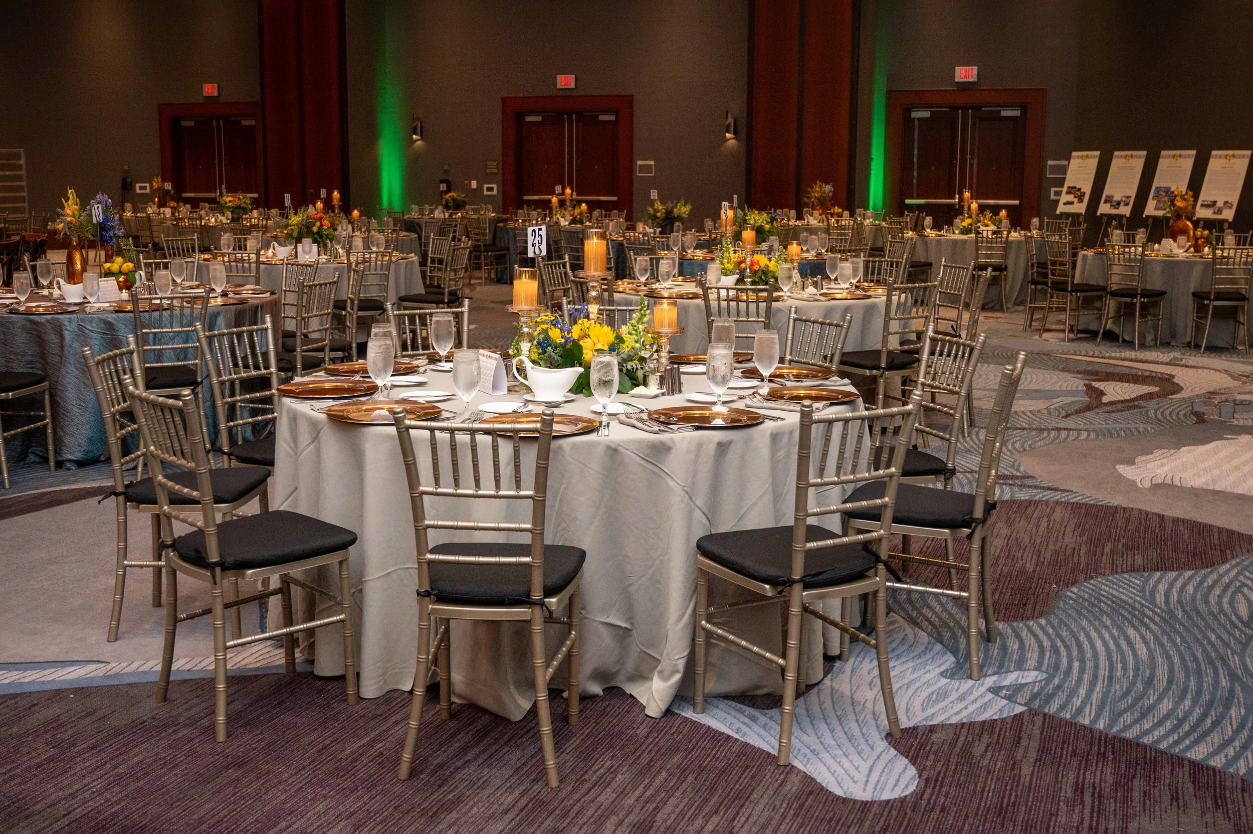 Event space with round tables covered in white tablecloths, decorated with floral centerpieces and candles, set for a formal occasion in a dimly lit room with dark walls and green accent lighting.