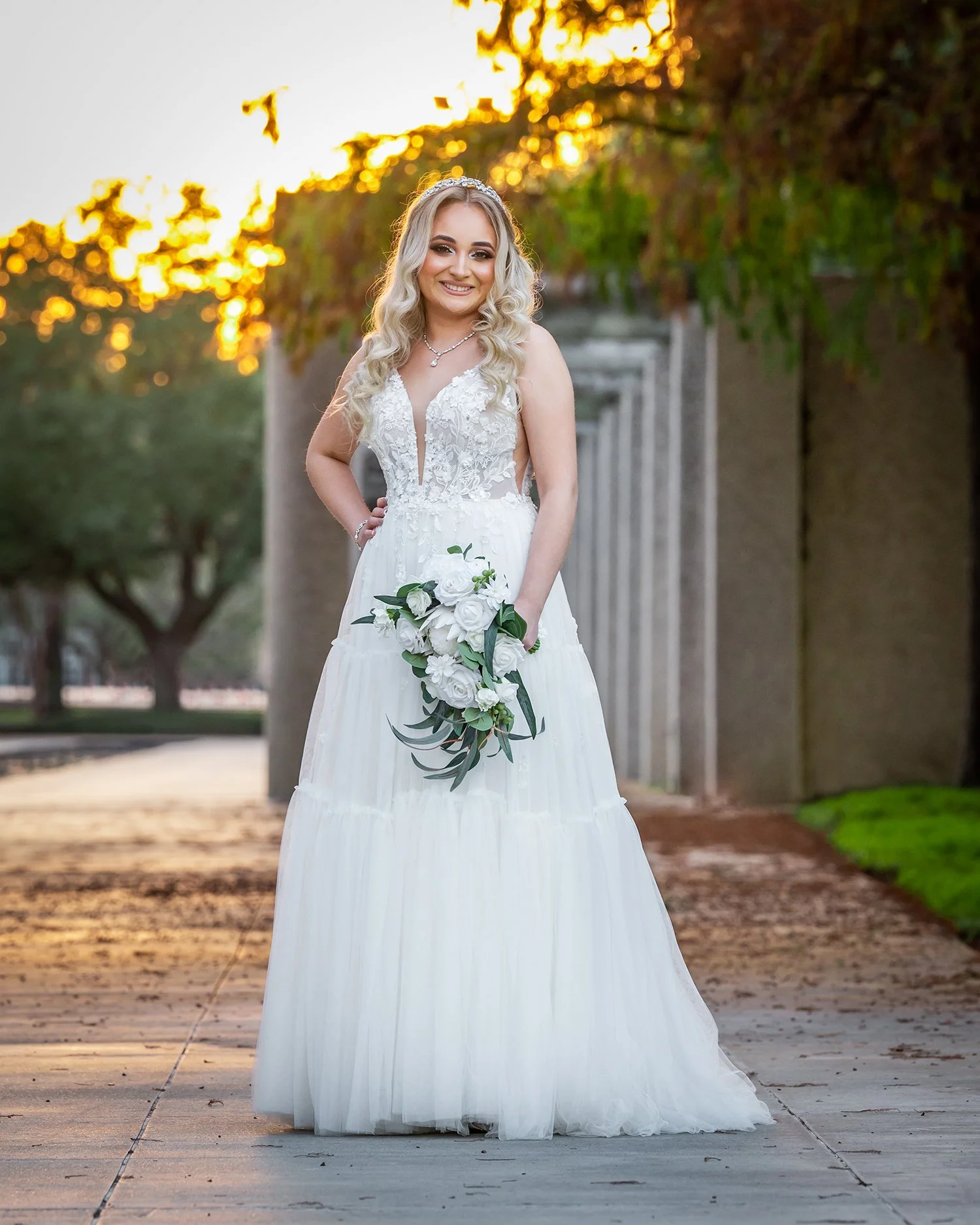 A young woman in a white wedding dress standing outdoors on a sidewalk during sunset, holding a bouquet of white flowers with green leaves, smiling at the camera.