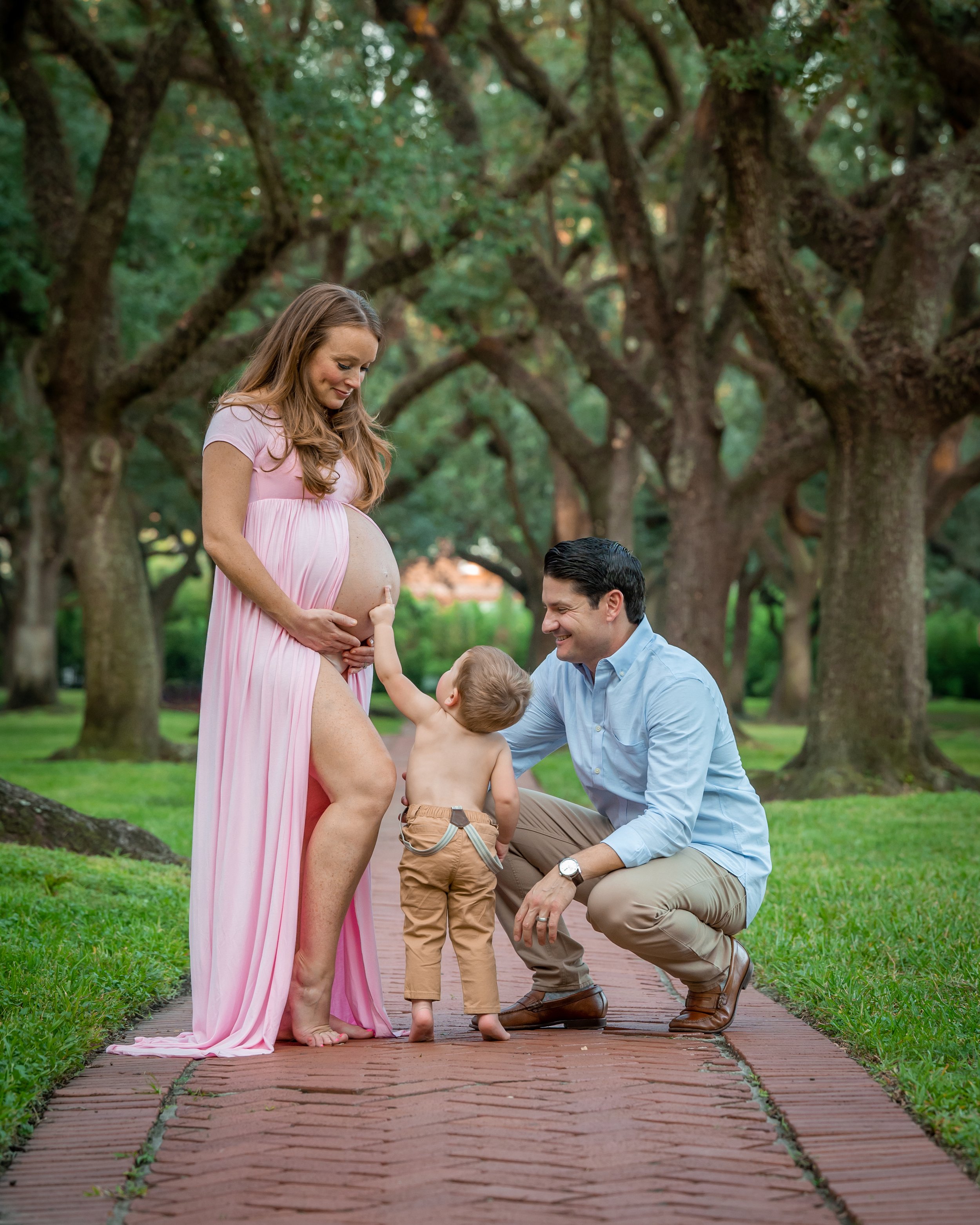 Pregnant woman in a pink dress with a child, an adult man kneeling and holding the child's hands, outdoors on a brick path with green grass and large trees in the background.