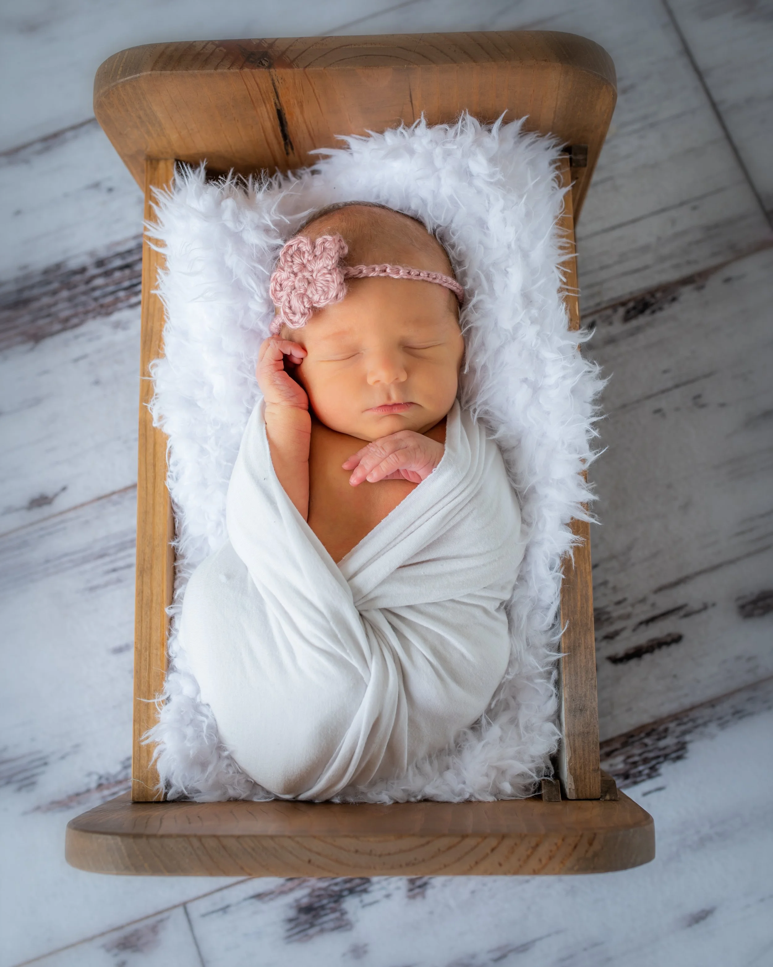 A sleeping newborn baby wrapped in a white blanket, lying on a fluffy white cushion on a wooden surface with a rustic, weathered finish.
