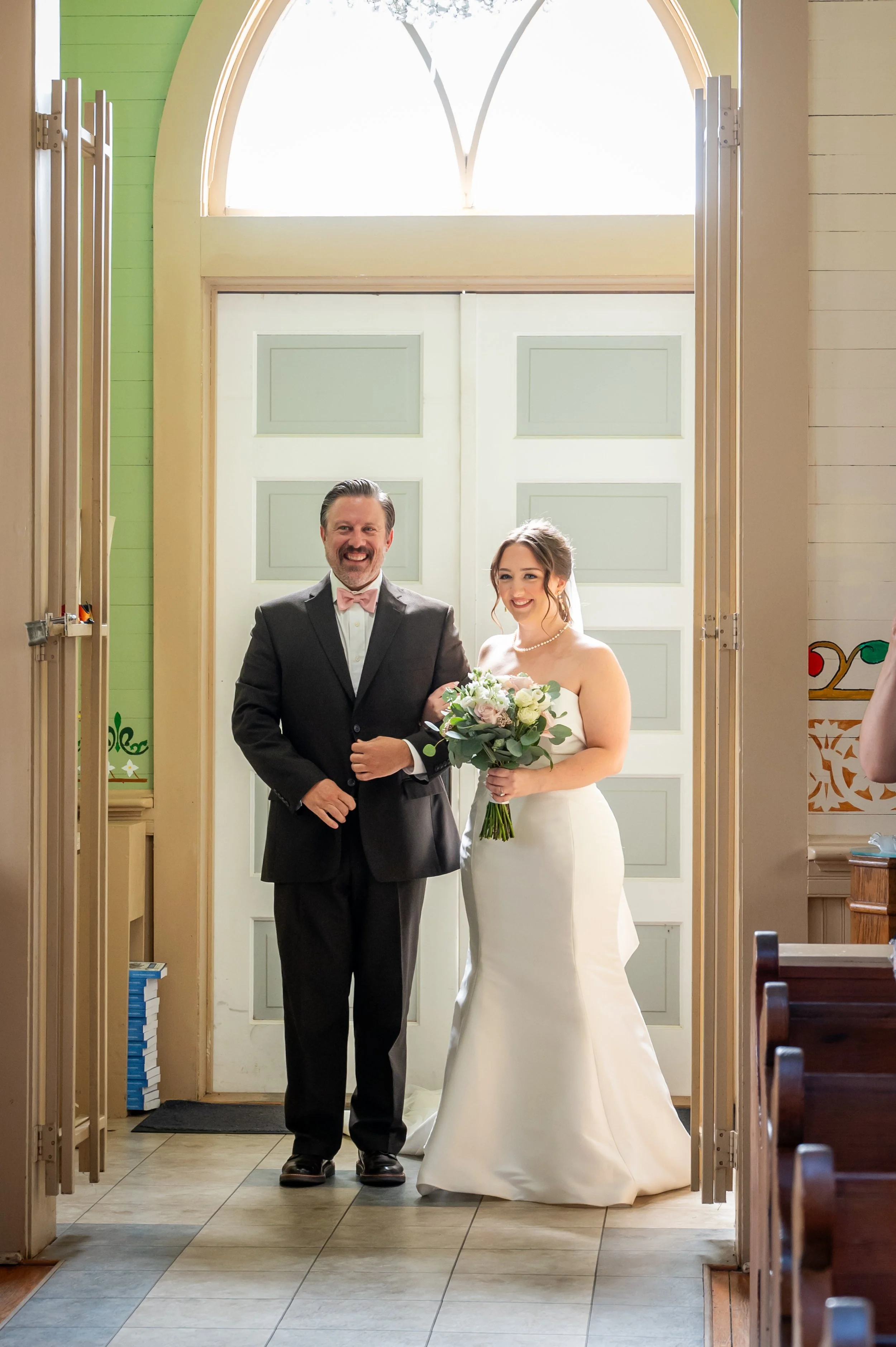 A father in a tuxedo with a pink bow tie and a woman in a white wedding dress holding a bouquet of flowers, standing inside a church doorway.