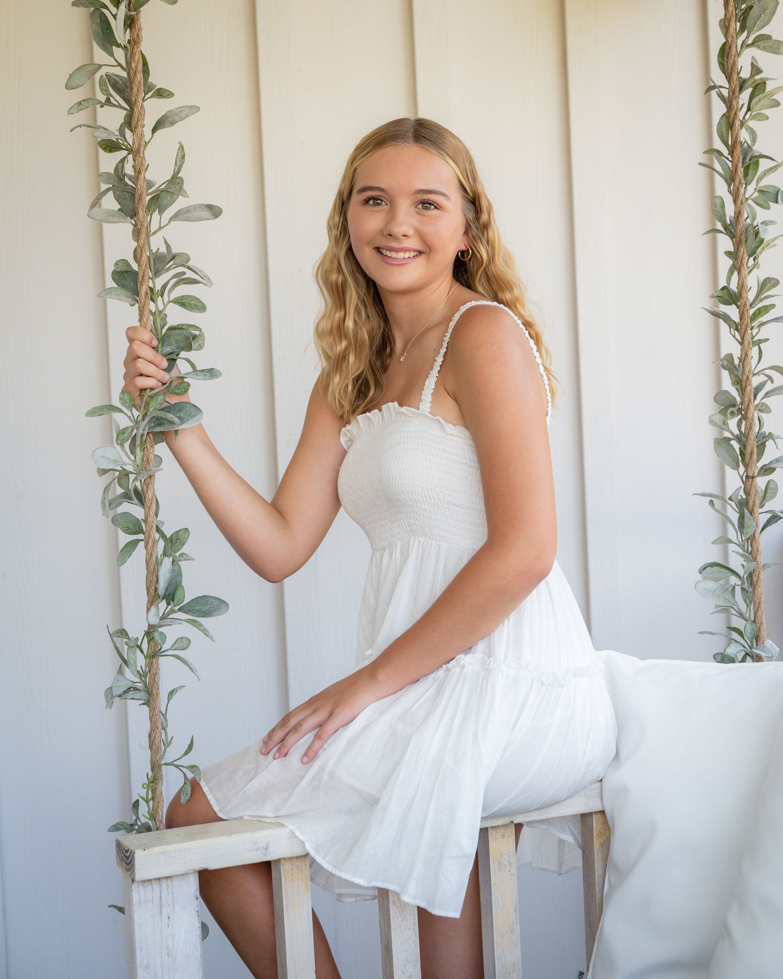 A senior girl with long, curly blonde hair smiling while sitting on a swing with rope handles and greenery on either side, in front of a cream-colored wall.