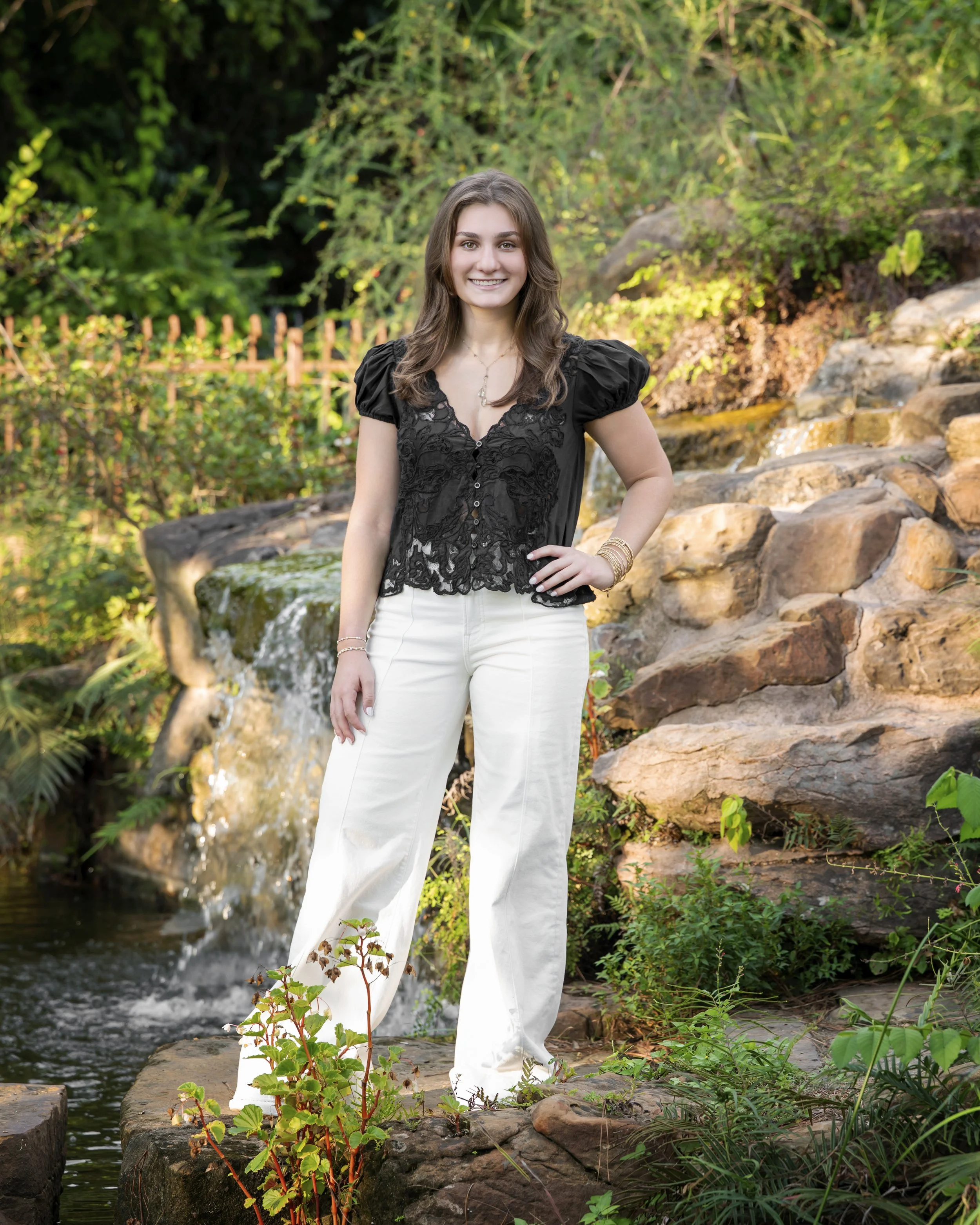 A young woman with long wavy brown hair standing near a small waterfall and rocks in a lush green natural setting, smiling at the camera.