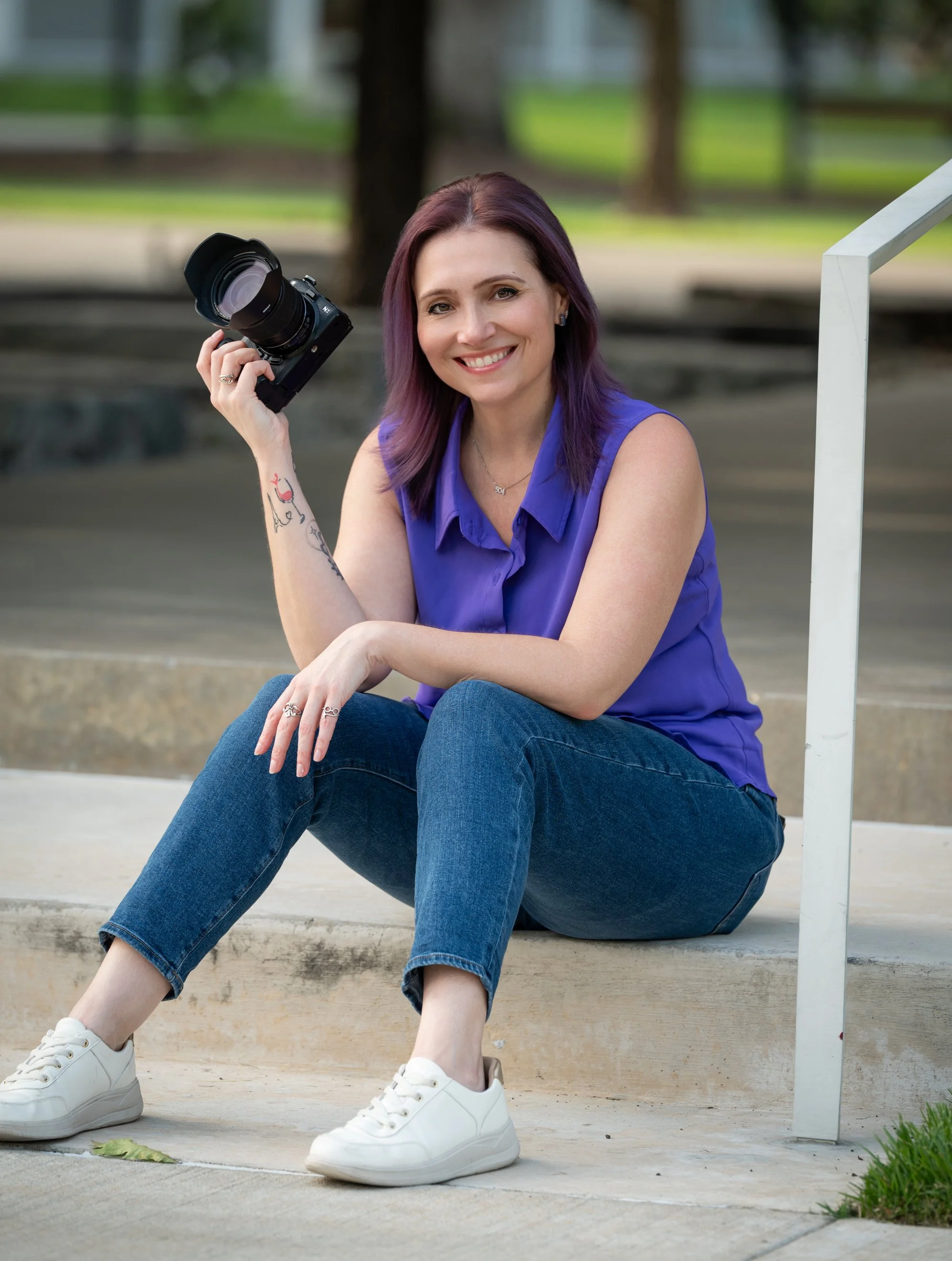 A woman photographer with purple hair and a blue sleeveless shirt sitting on concrete steps outdoors, holding a camera with a large lens, smiling at the camera.