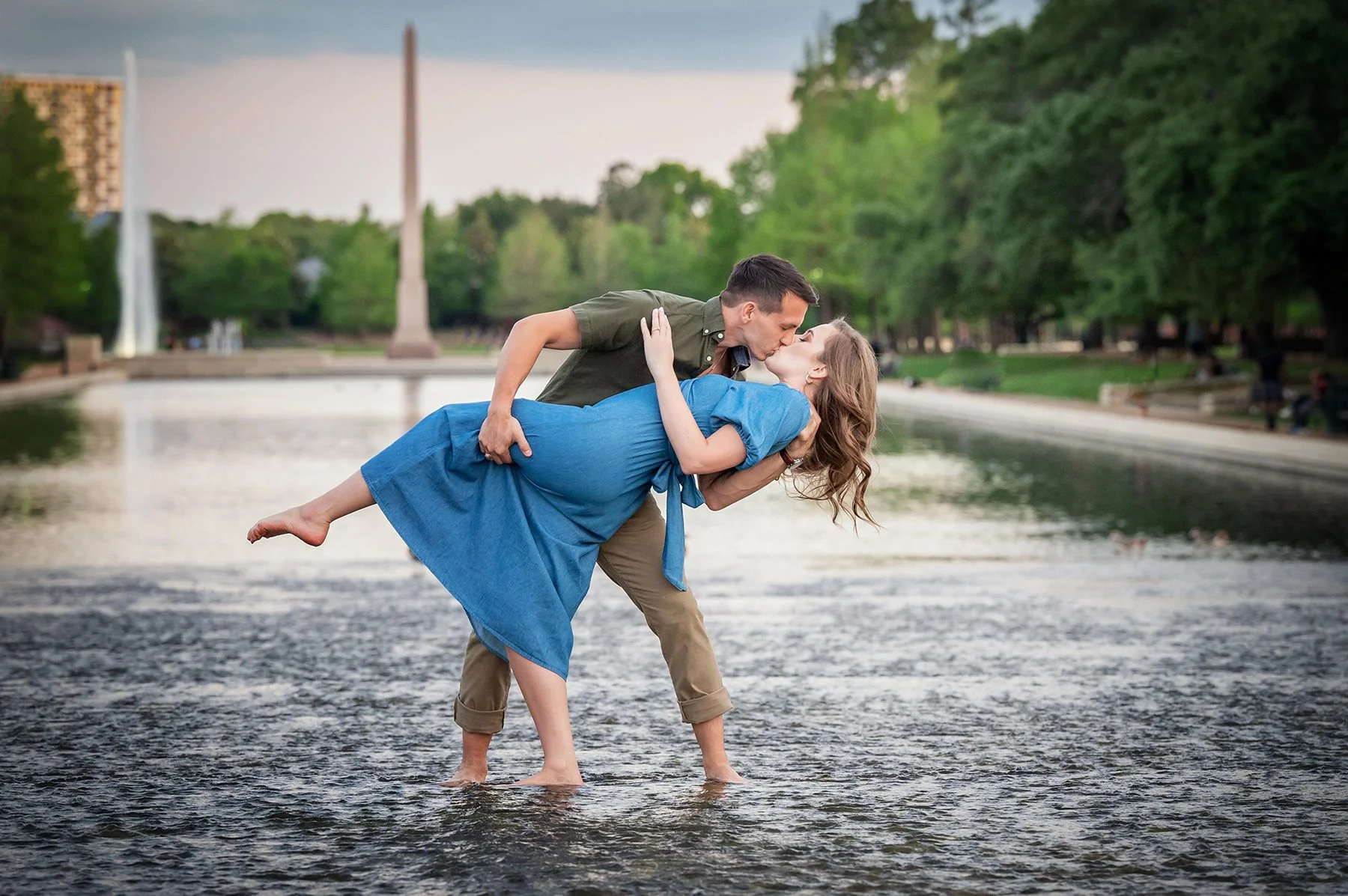 A man in khaki pants and a woman in a blue dress kiss as he dips her in a shallow river in a park with trees, a water fountain, and a monument in the background.