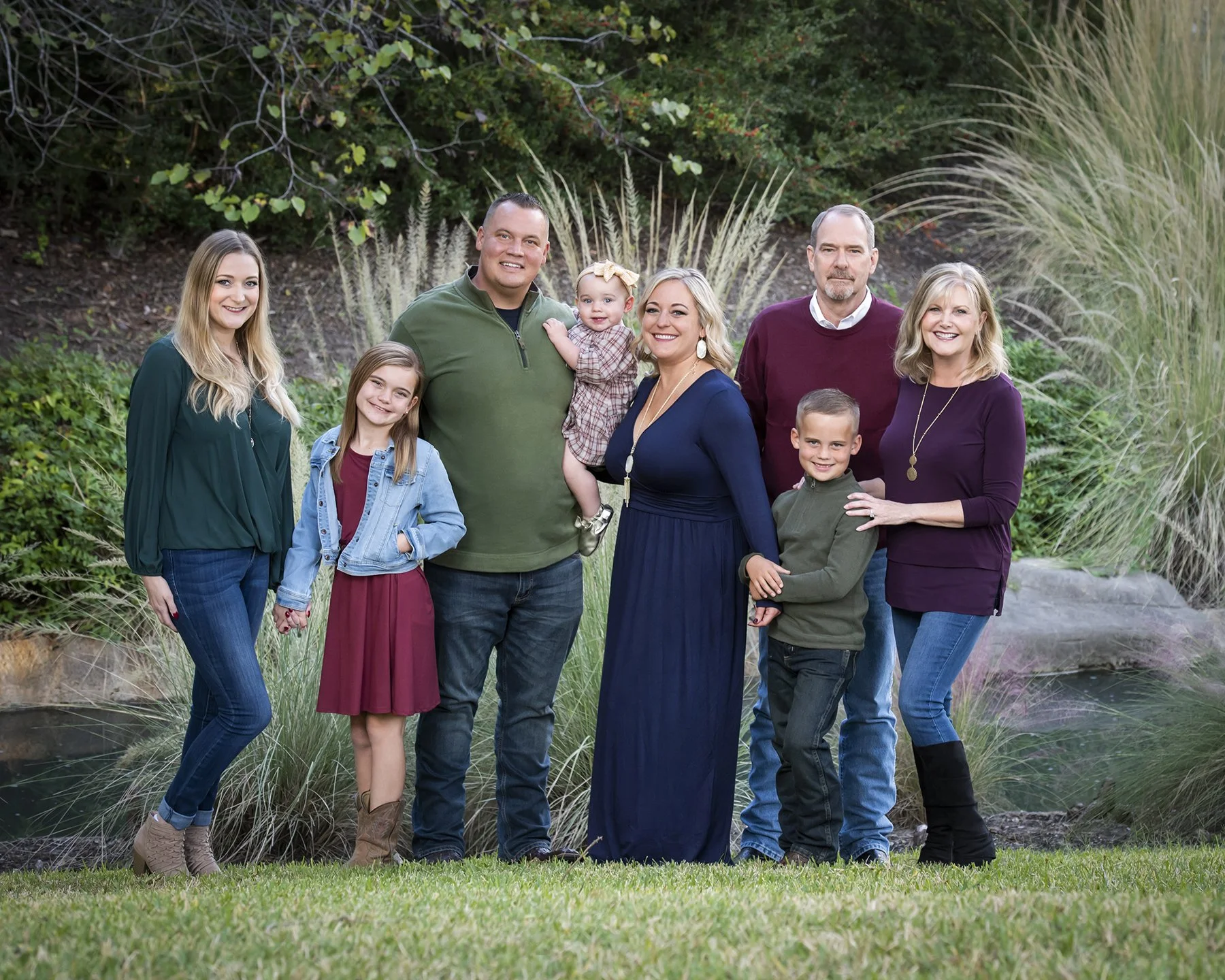 Family group standing outdoors on grass with bushes and trees in the background, smiling at the camera.