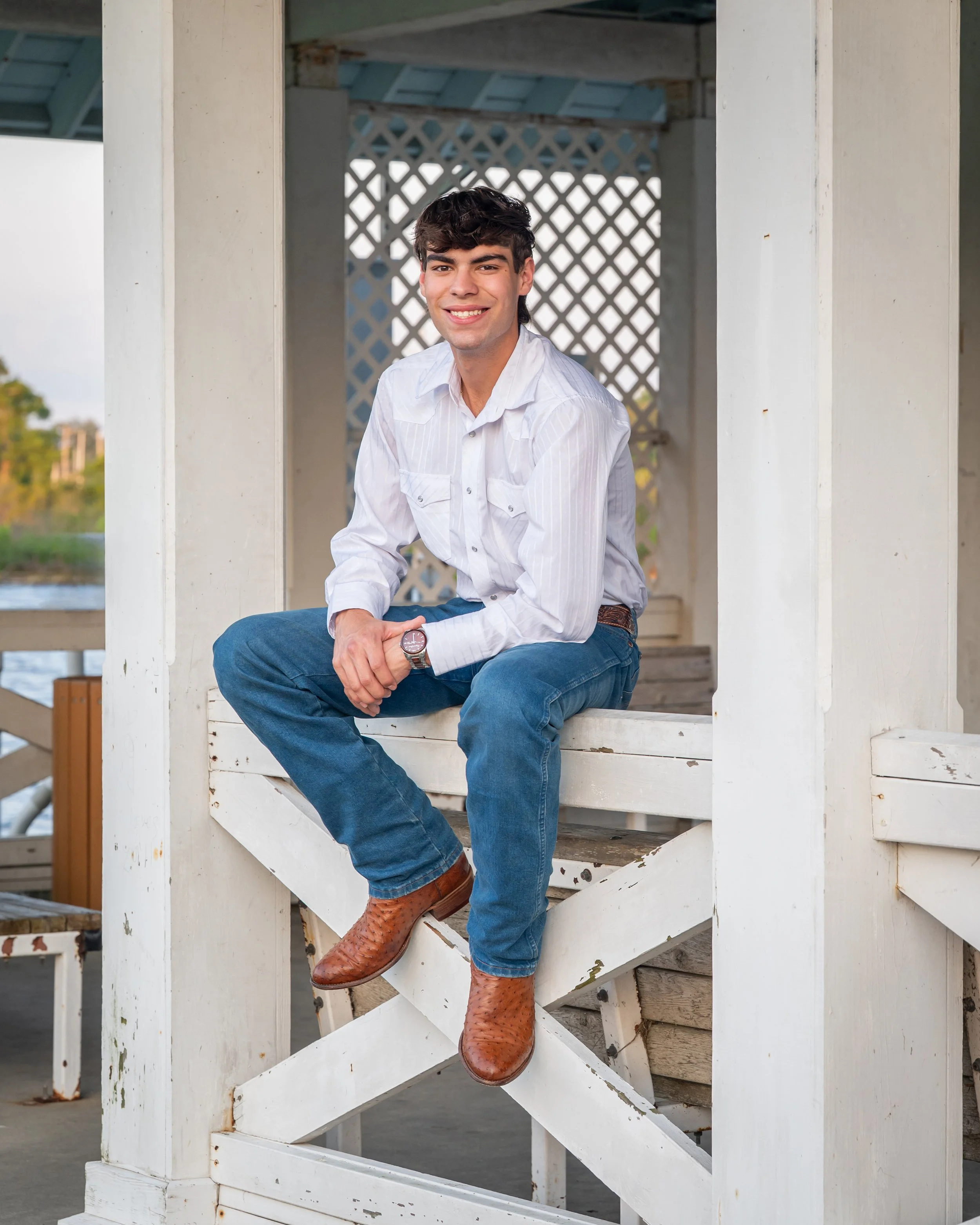 A senior guy wearing a white long-sleeve button-up shirt, blue jeans, and brown cowboy boots sitting on white wooden stairs in a gazebo with a lattice back and railing.