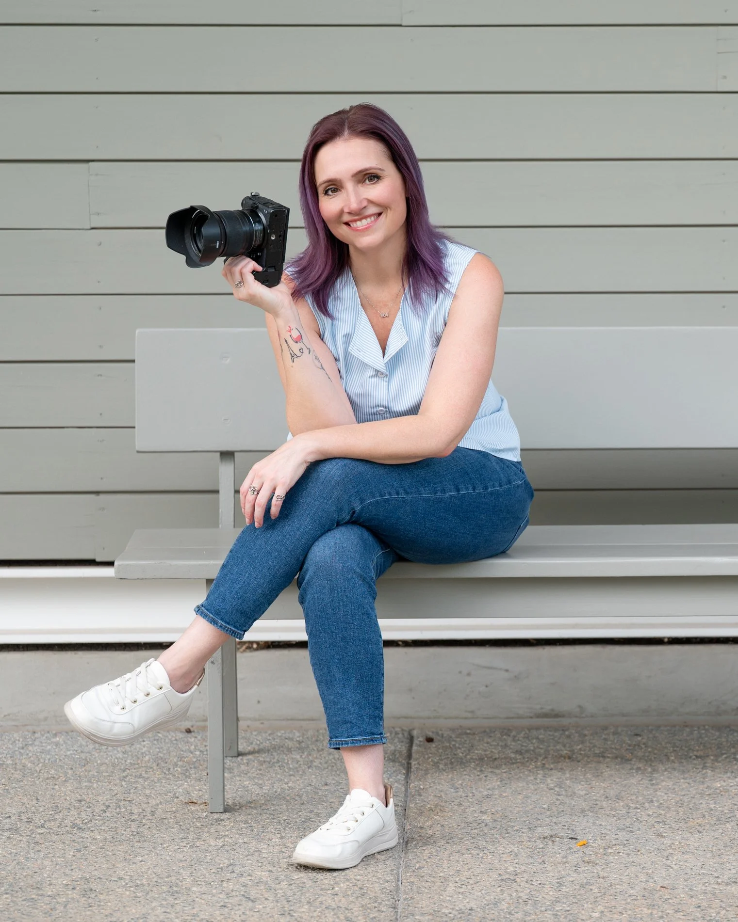 A woman photographer with purple hair sitting on a gray bench against a gray wooden wall, holding a camera, wearing a sleeveless striped shirt, blue jeans, white sneakers, and smiling.