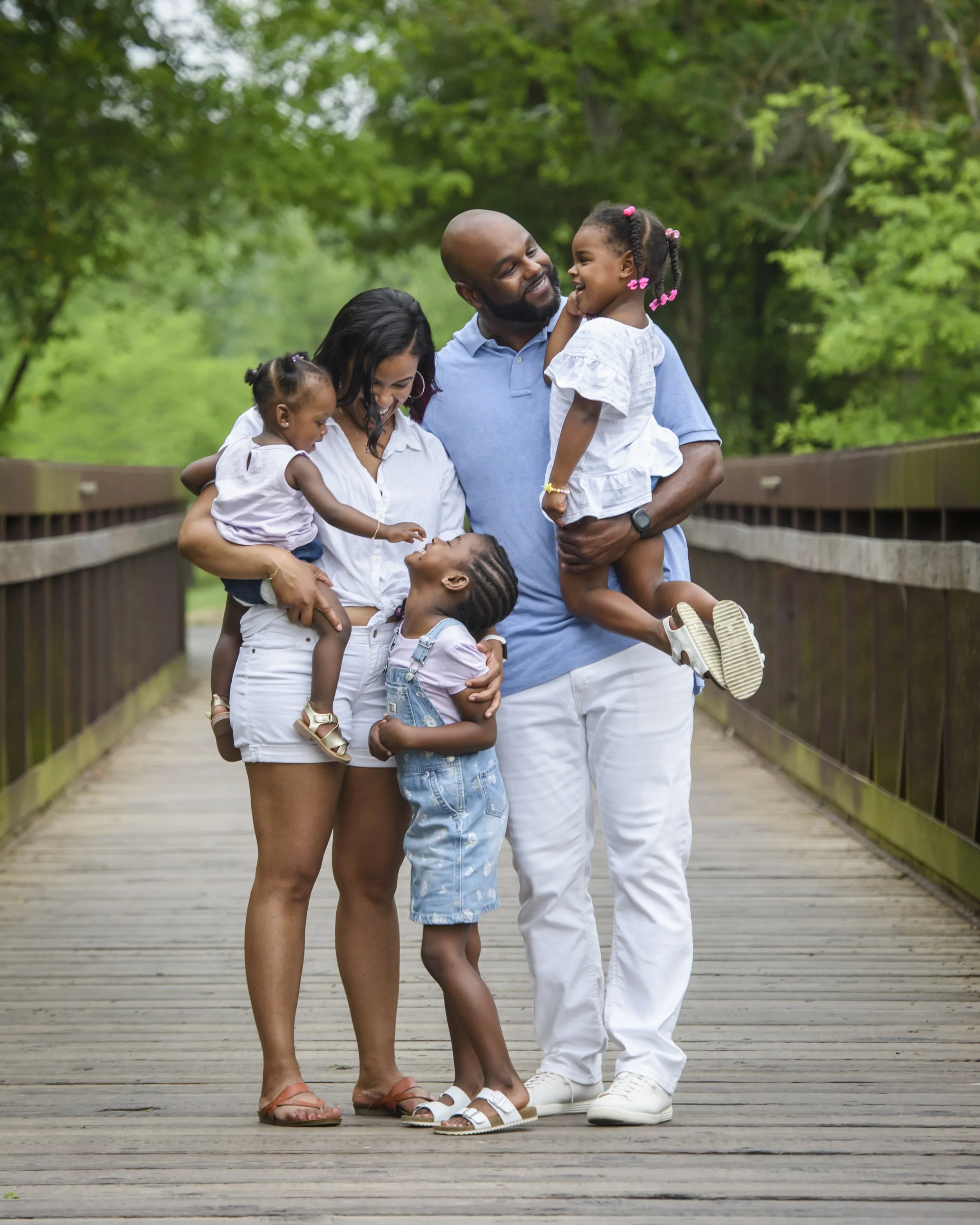 A family of five, including two young children, enjoying a walk on a wooden bridge in a lush green park, smiling and laughing together.