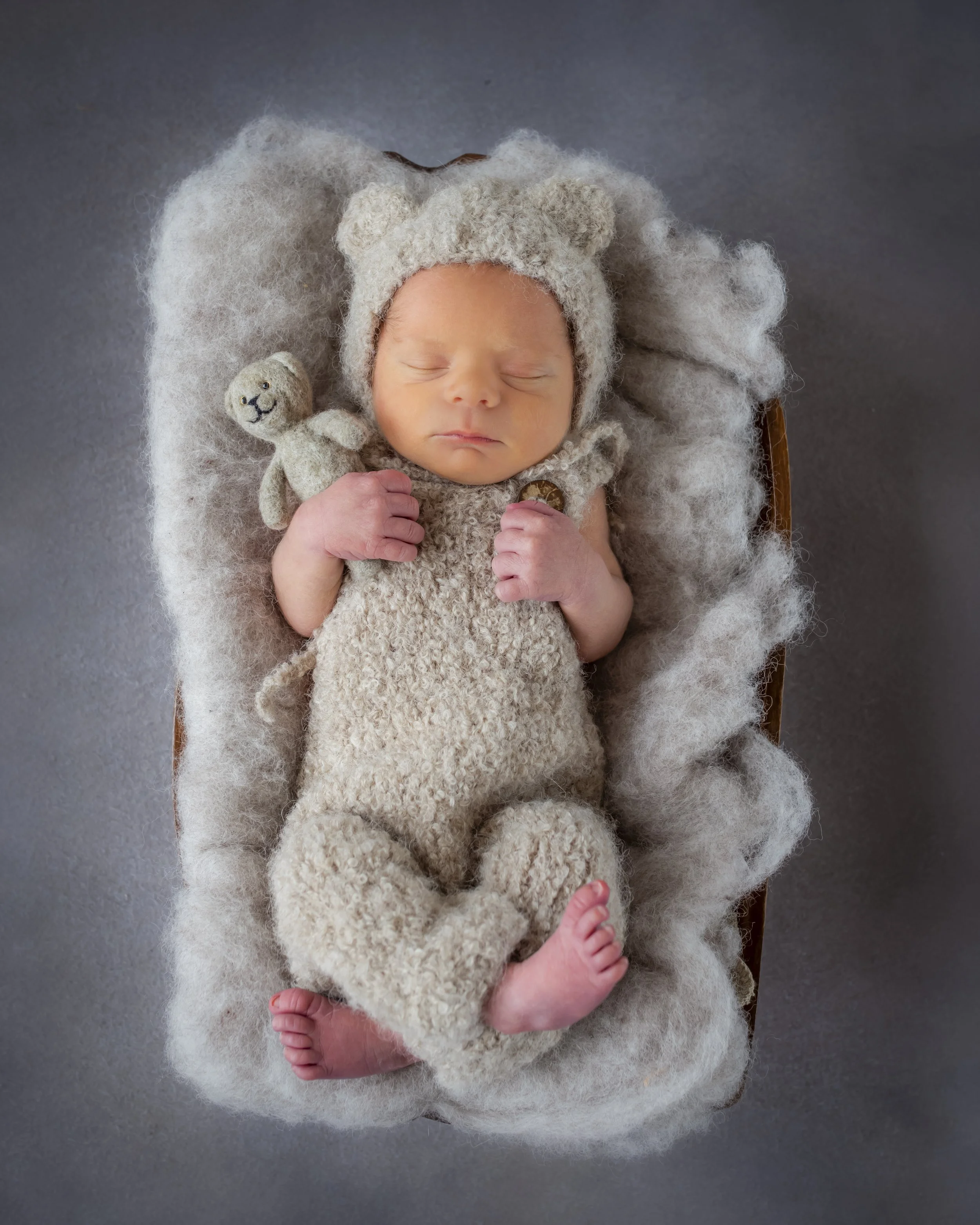 A newborn baby dressed in a cozy, knitted bear costume with a bear face hat. The baby holds a small stuffed bear toy while lying on a fuzzy blanket.