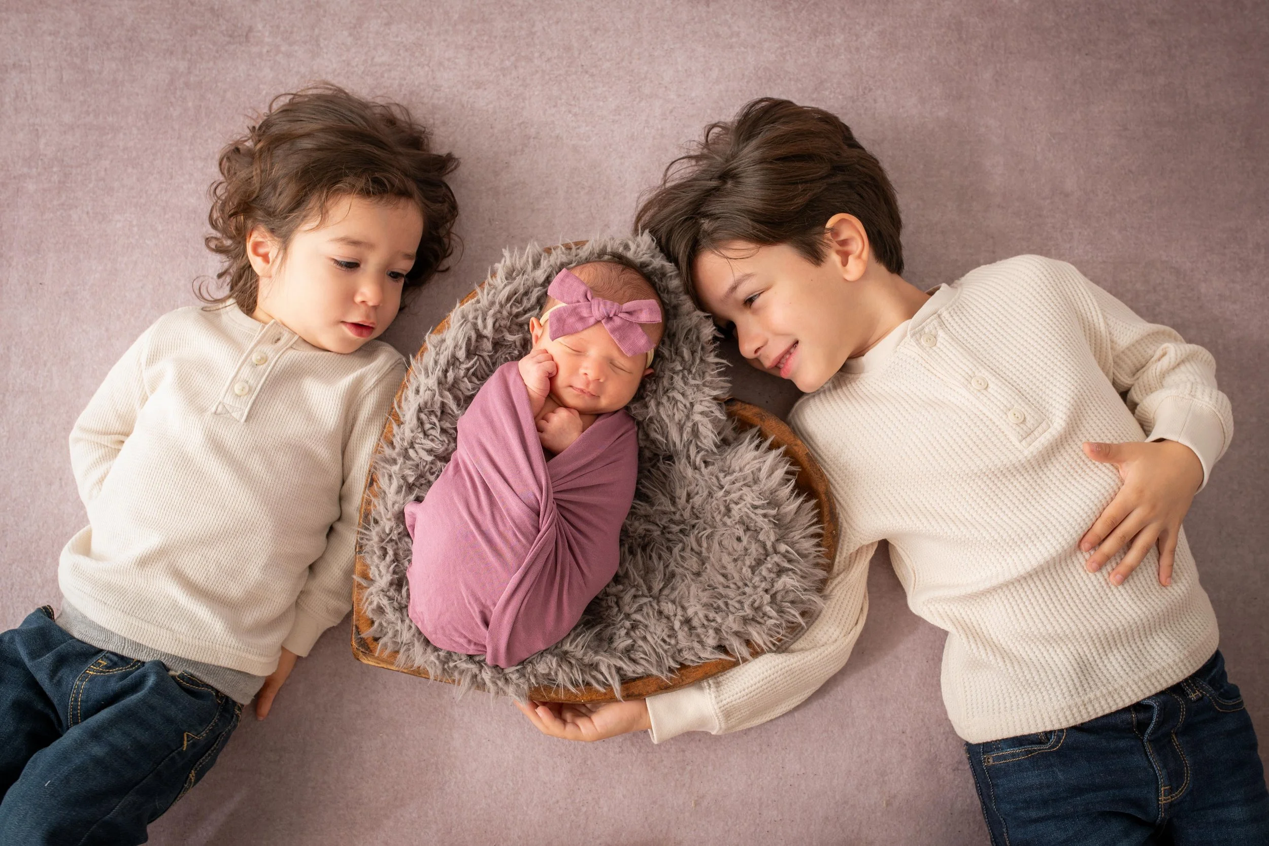 Three children lying on a pink carpet, surrounding a baby in a basket with a furry gray blanket. The newborn baby has a purple headband with a bow and is asleep.