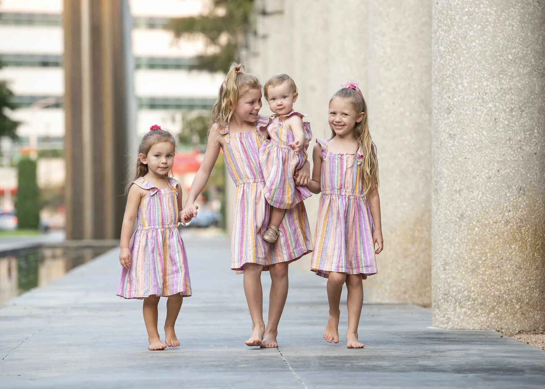 Four young girls walking together outdoors holding hands; three of them are older, and one is a baby being carried by one of the older girls. All girls are smiling, and they are dressed in matching pink and purple striped dresses.