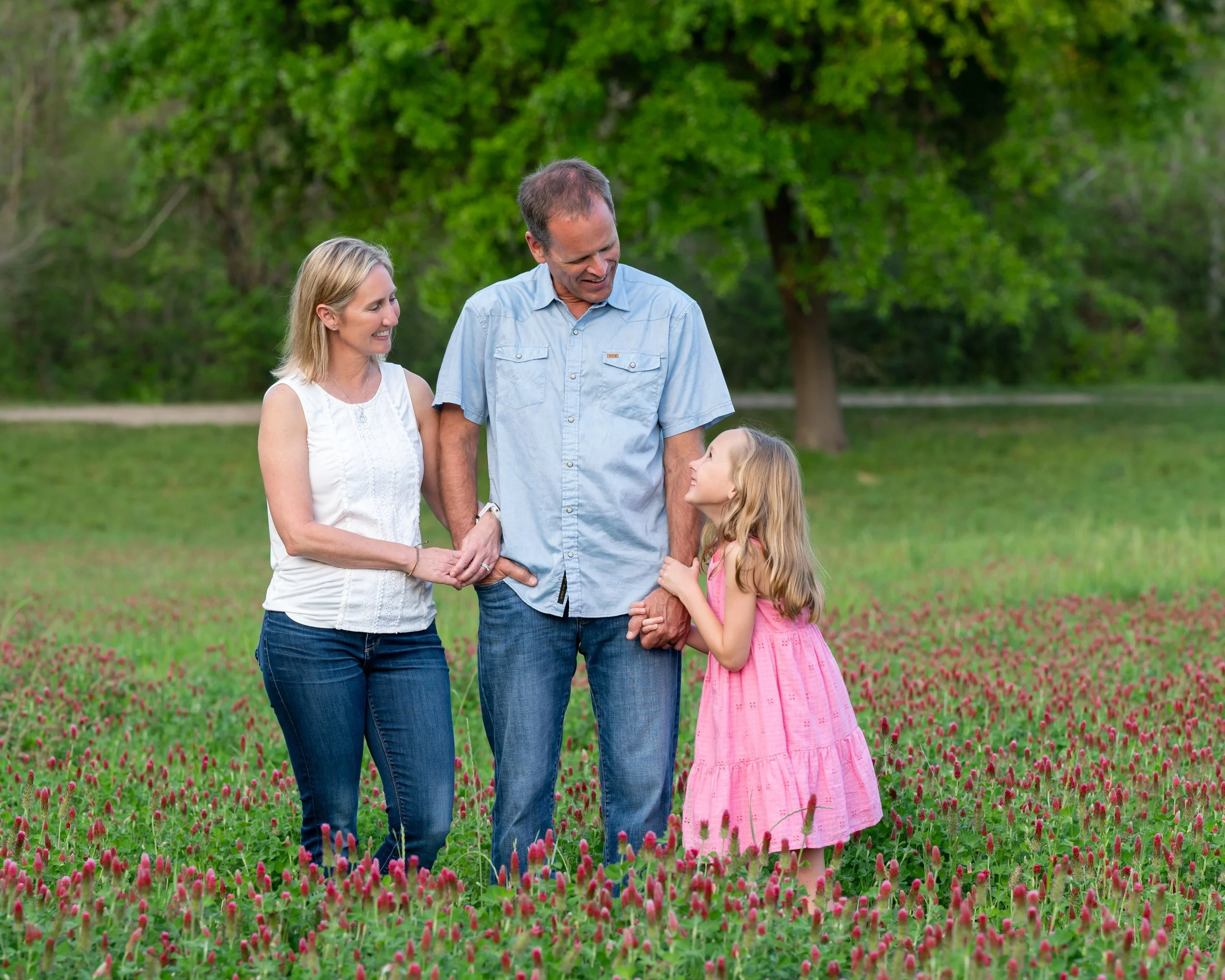 A family of three, including a woman, a man, and young girl, standing together outdoors in a lush green park with pink flowers in the foreground, smiling and looking at each other.