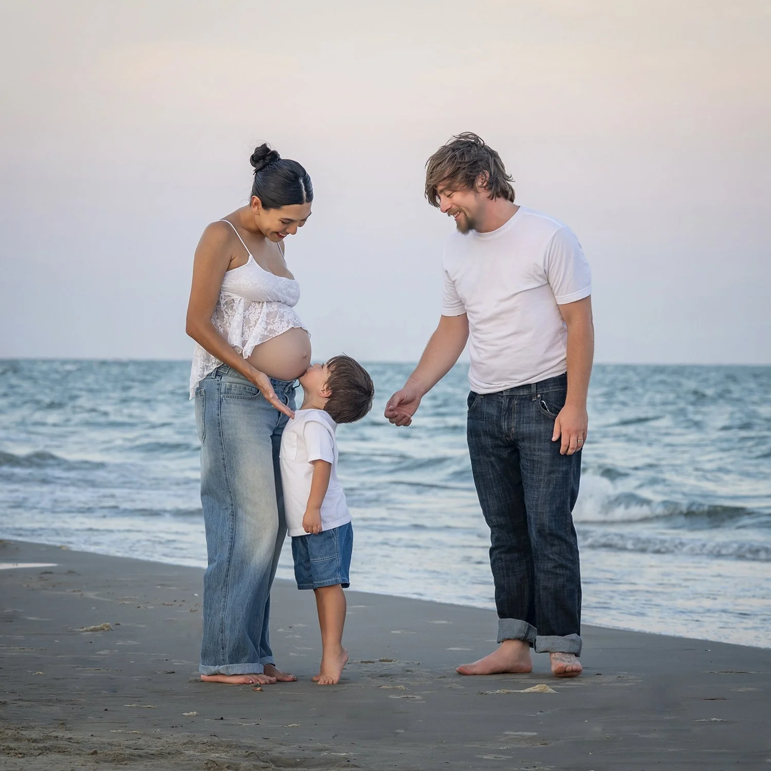 A family of three at Galveston beach, with a pregnant woman, a man, and a child, smiling and holding hands, near the ocean at sunset.