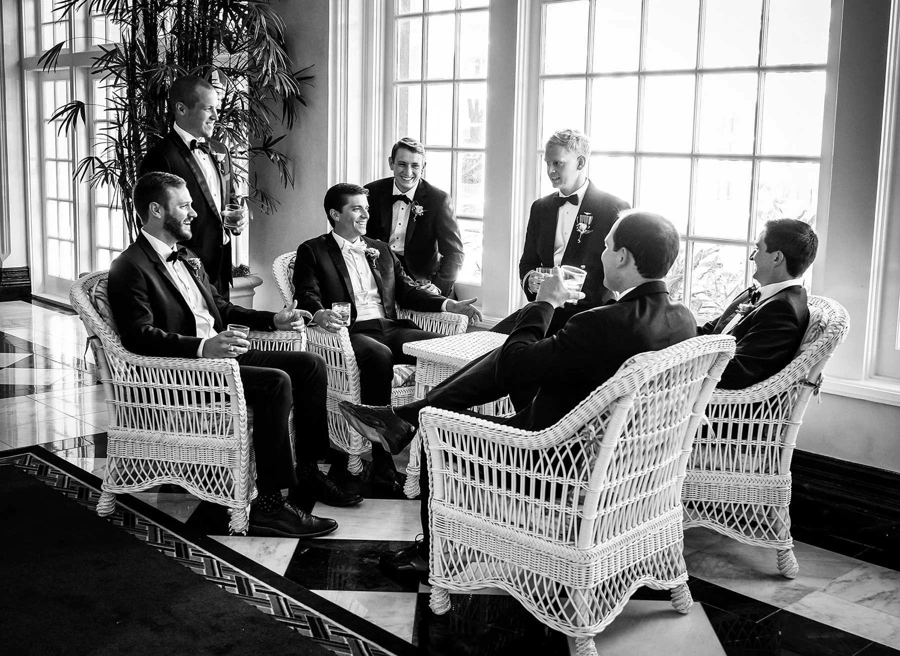 A group of seven men in formal tuxedos, some with medals, sitting and standing around wicker chairs with drinks in a bright room with large windows, greenery, and tiled floors, engaging in conversation.