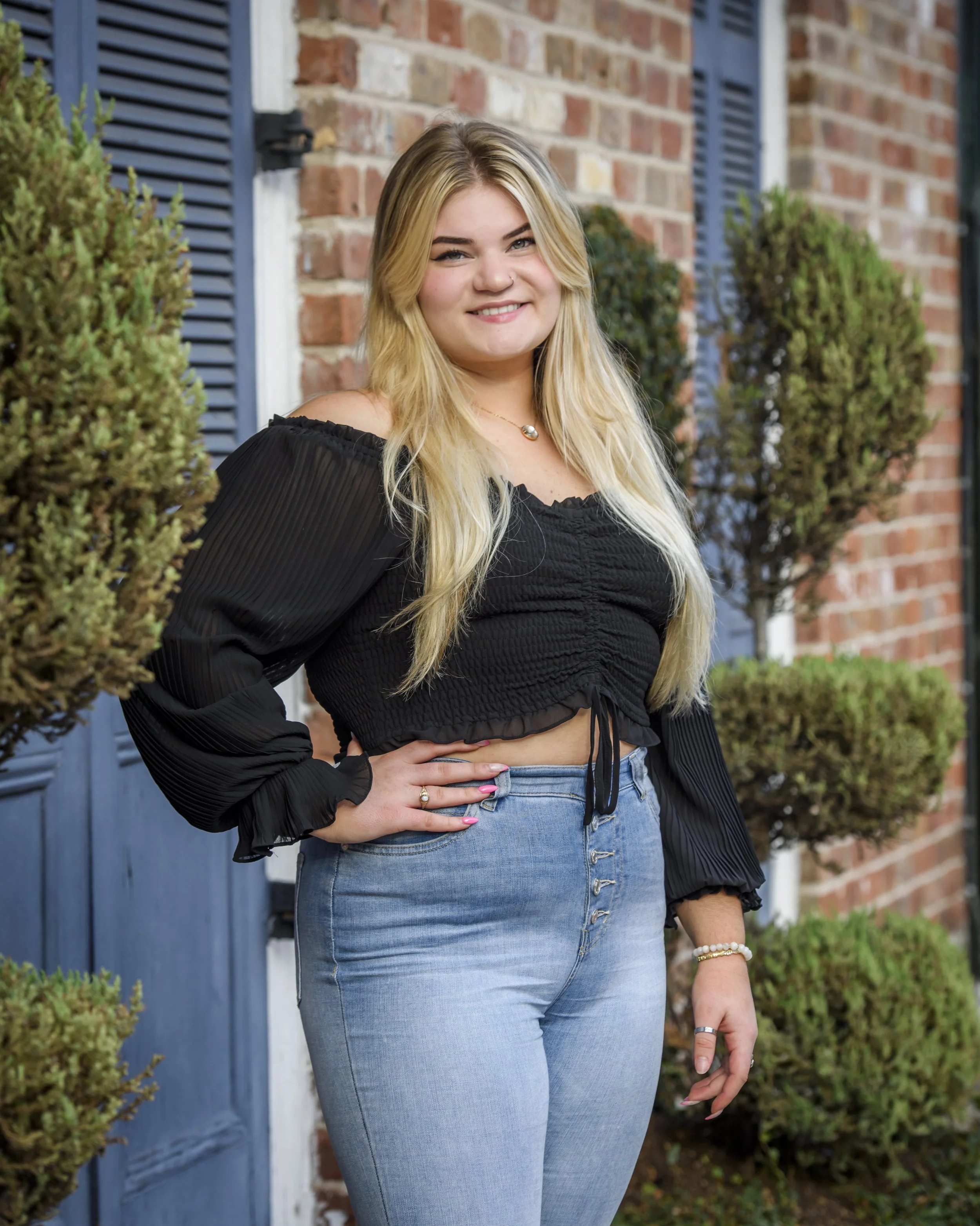 senior girl  with long blonde hair wearing a black off-shoulder top and blue jeans, standing outside near a brick wall with a blue door and greenery.