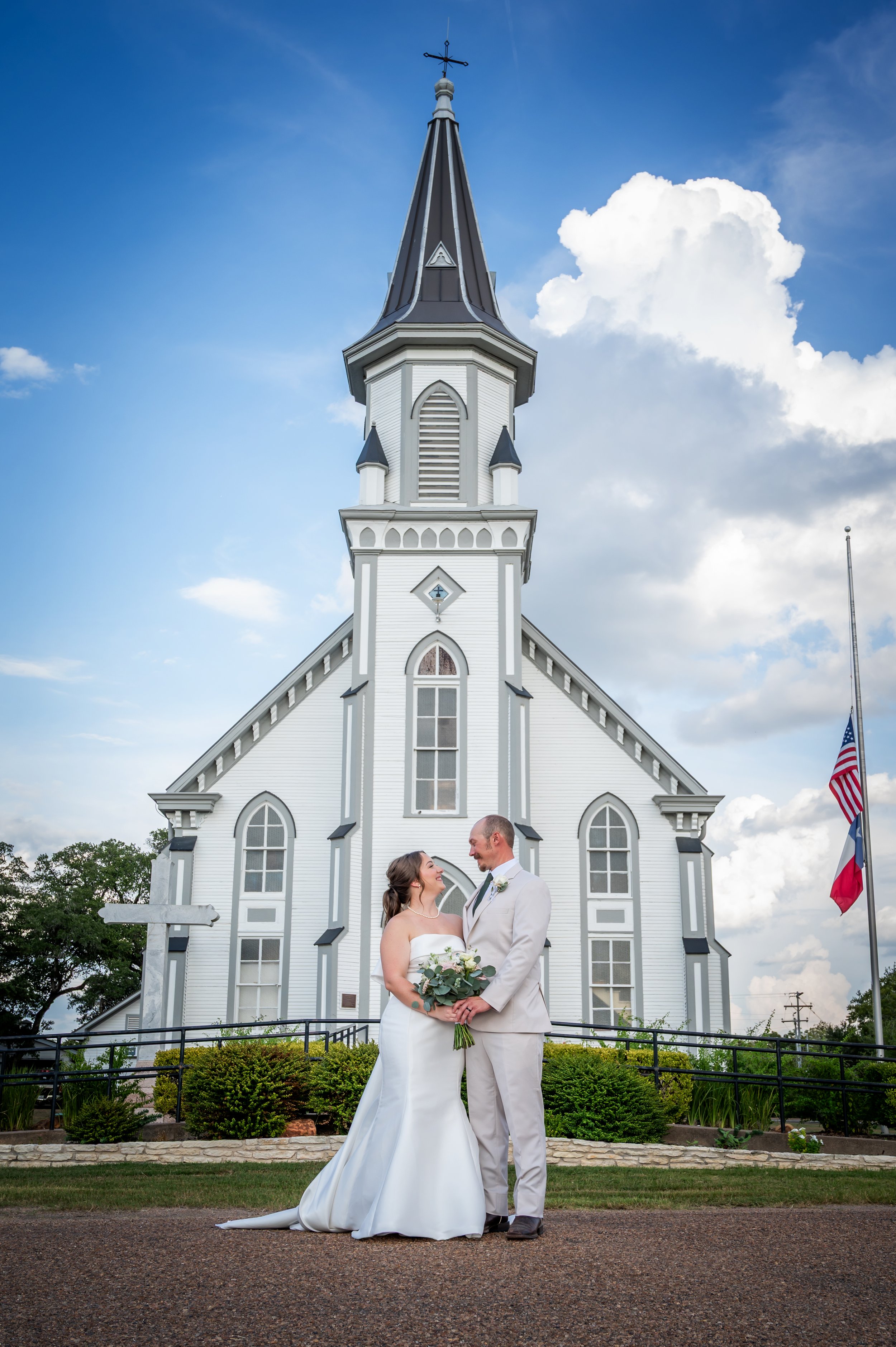 A bride and groom in wedding attire standing in front of a white church with a tall steeple and cross, holding a bouquet with a cloudy sky in the background.