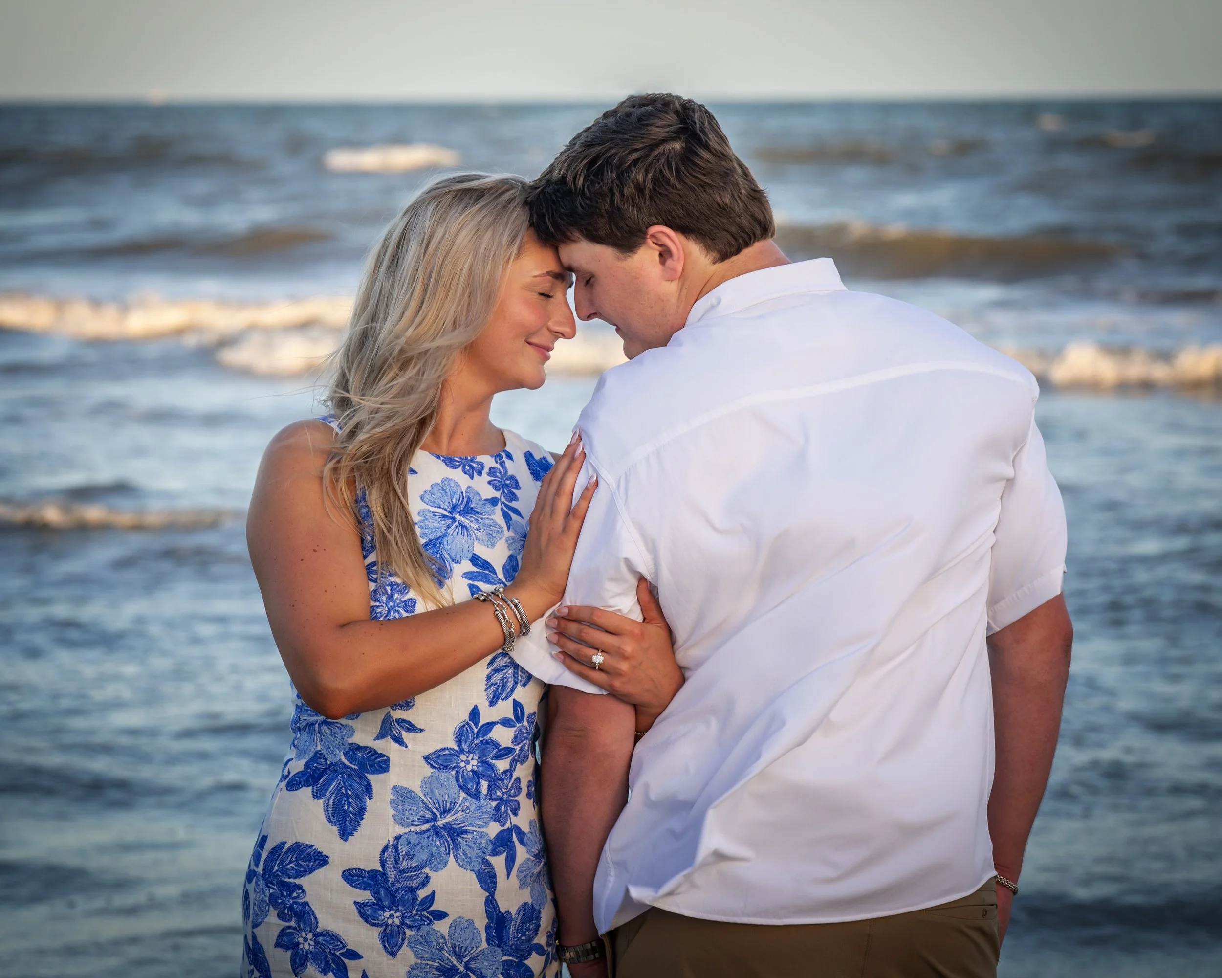 A couple stands close at Galves beach, with their foreheads touching, eyes closed, and gentle smiles. The woman has long blonde hair and is wearing a white dress with blue floral patterns. The man has short brown hair, a white shirt, and khaki pants.
