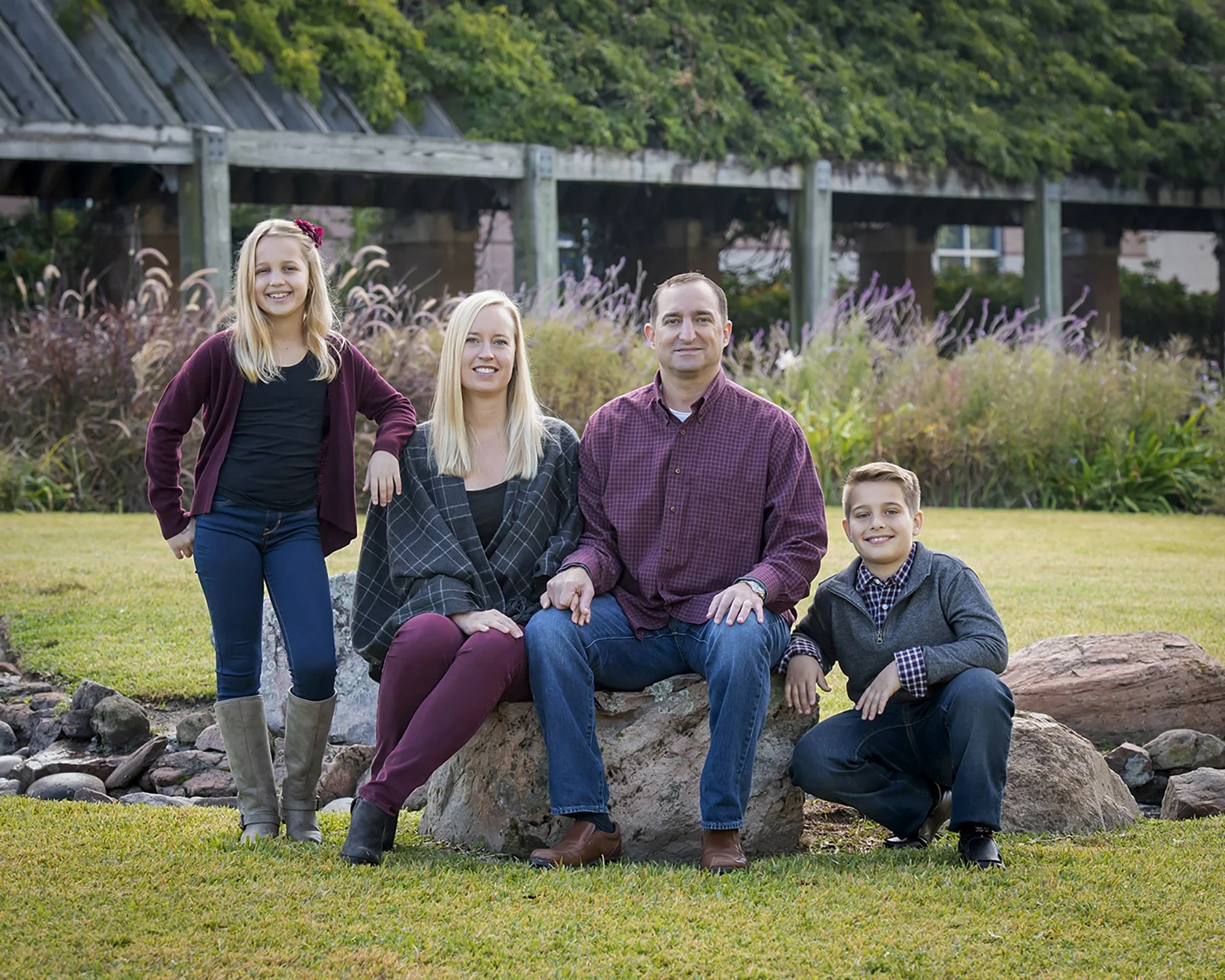 A family of four posing outdoors on a grassy area with rocks, plants, trees, and a wooden bridge or structure in the background. The family includes a girl, a woman, a man, and two boys, all smiling at the camera.