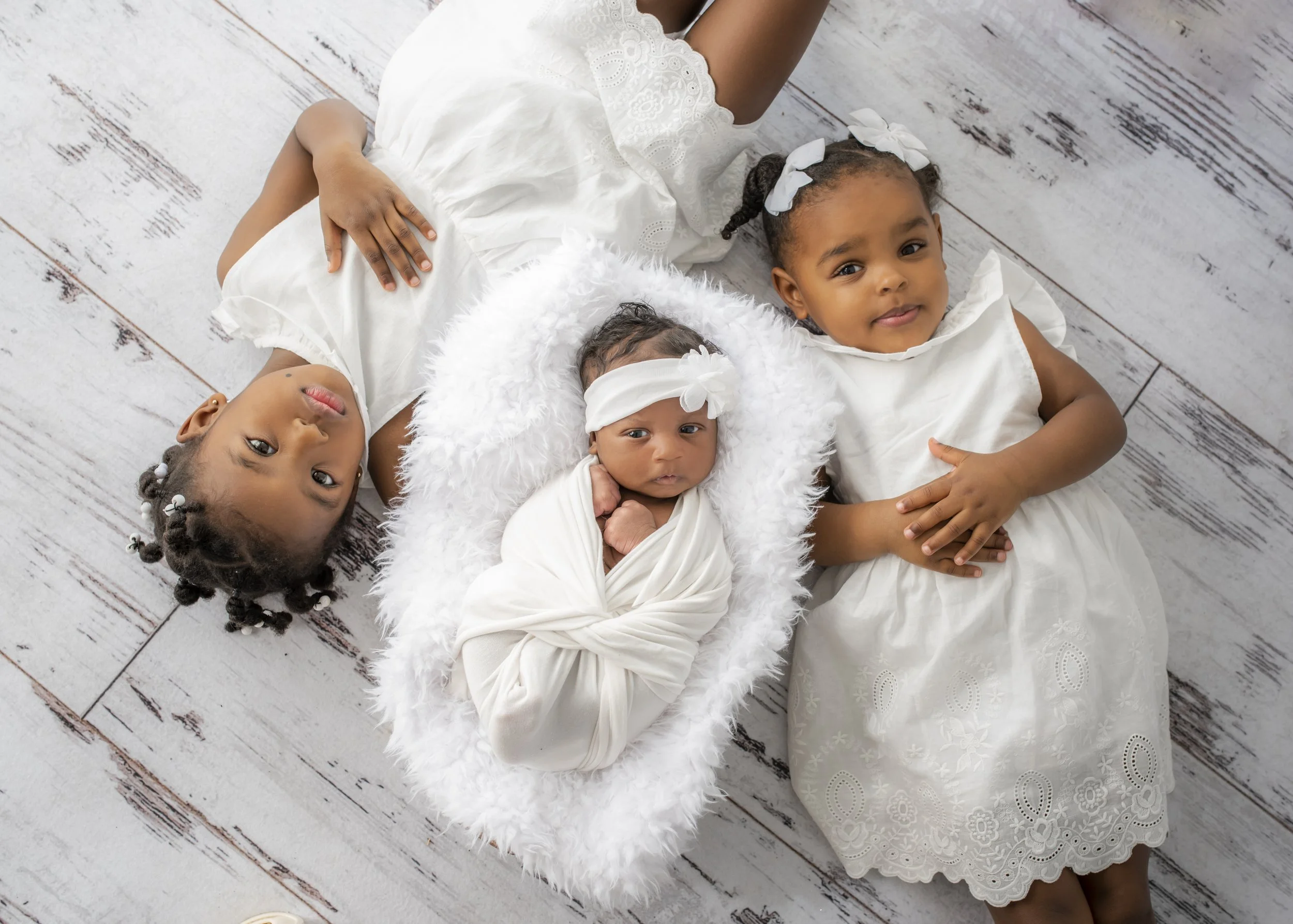 Three young girls lying on a wooden floor, dressed in white dresses, one of them is a newborn with a headband, and the other two are older with bows in their hair.