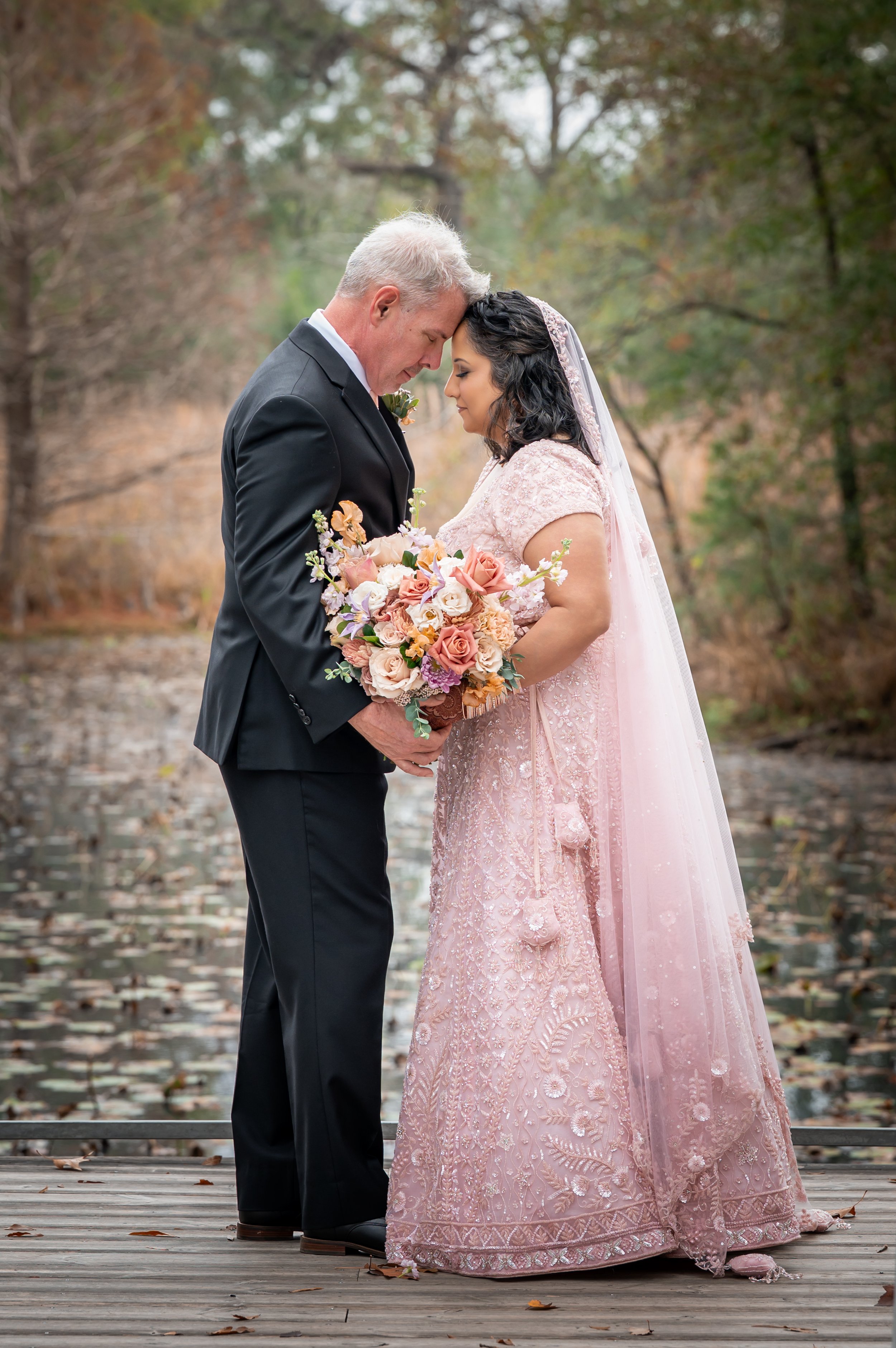 A couple dressed in wedding attire standing on a wooden dock with trees and water in the background, their foreheads touching and holding a bouquet of flowers.