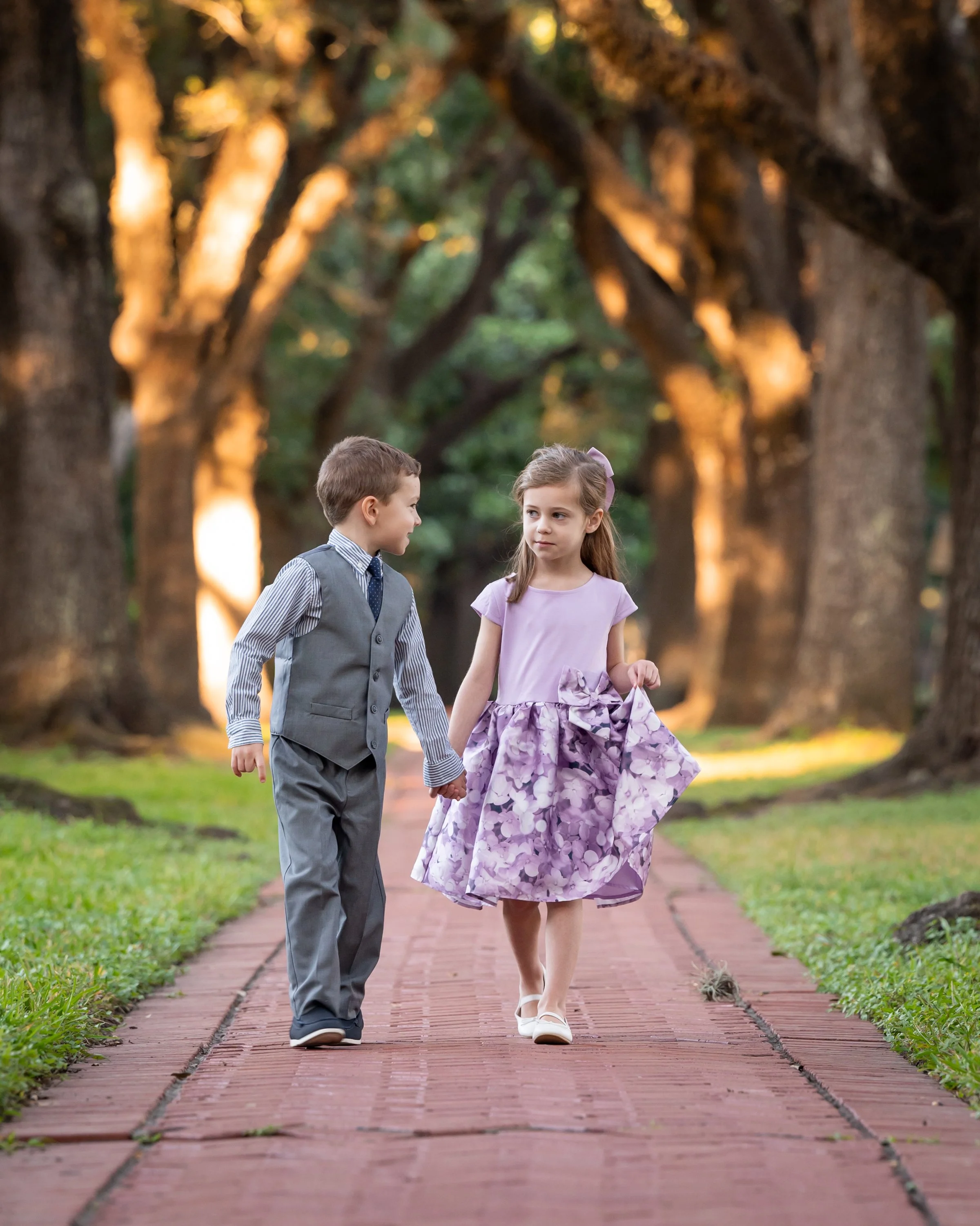 Two children, a boy and a girl, walking hand in hand on a brick pathway through a park with large trees at sunset.