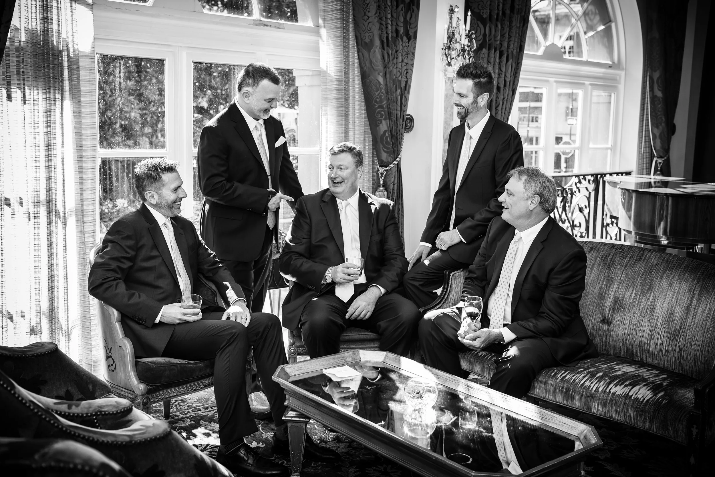 A group of six men dressed in suits is gathered in a well-lit room with large windows. They are smiling and laughing, with some seated on a vintage-style sofa and chairs, while others stand. One man in the center is holding a drink and appears to be 