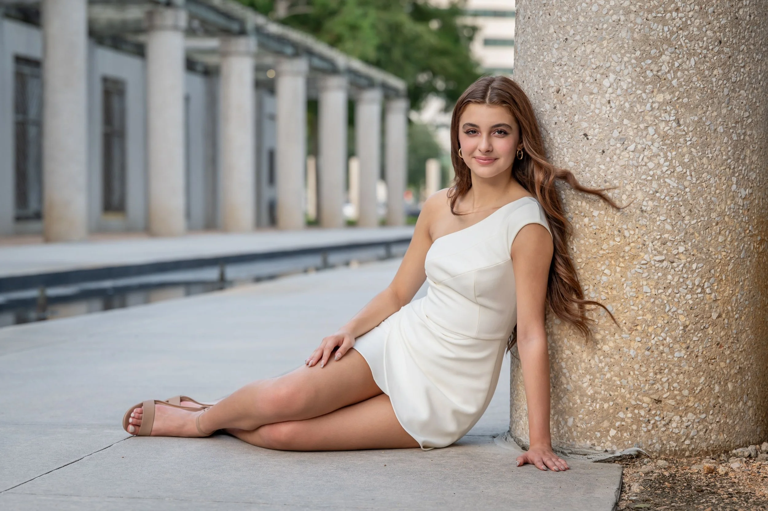 A young woman in a white dress is sitting on the sidewalk, leaning against a textured concrete pillar outdoors. She has long, wavy brown hair, and is wearing gold hoop earrings and beige sandals. The background features a modern building with tall columns and greenery.