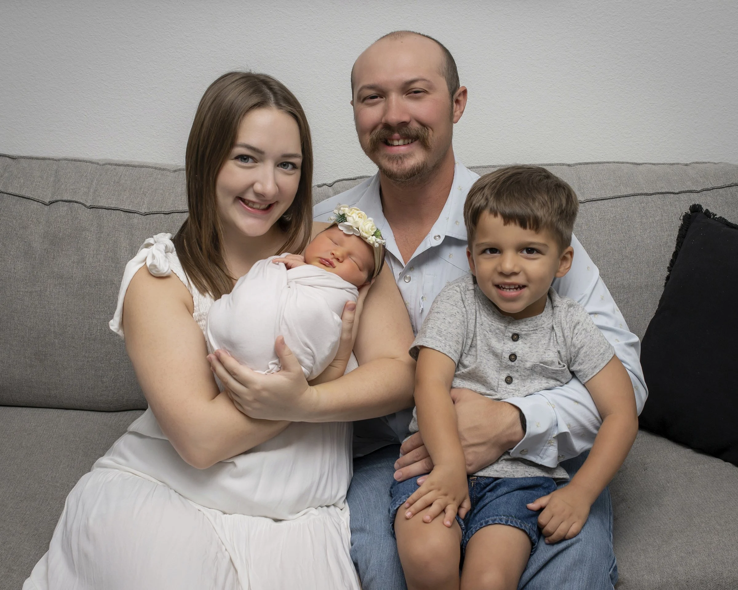 A family  sitting on a gray sofa against a white textured wall, including a woman with long brown hair holding a newborn baby with a floral headband, a man with a beard and mustache, a young boy with curly hair, and part of another person visible on 