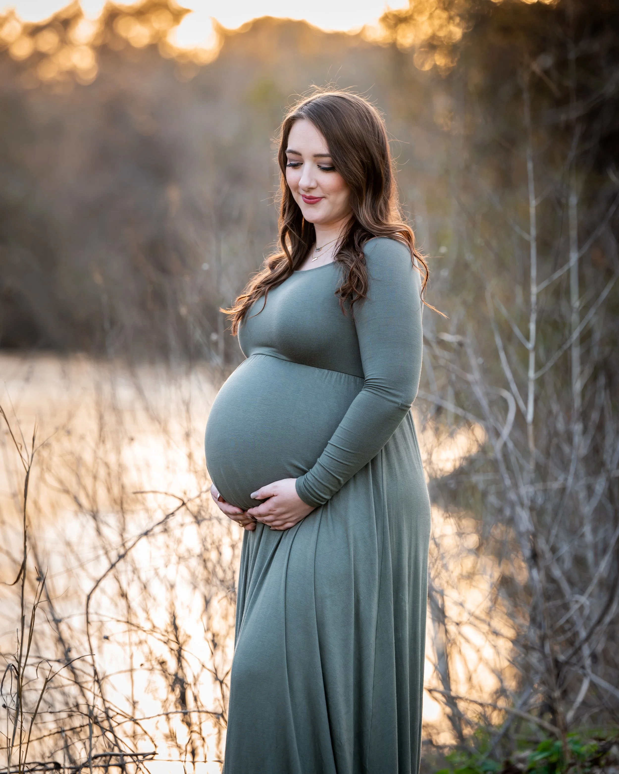 A pregnant woman with brown hair and makeup, wearing a long-sleeved, emerald dress, standing outdoors in a natural setting during sunset, gently holding her belly and smiling softly.