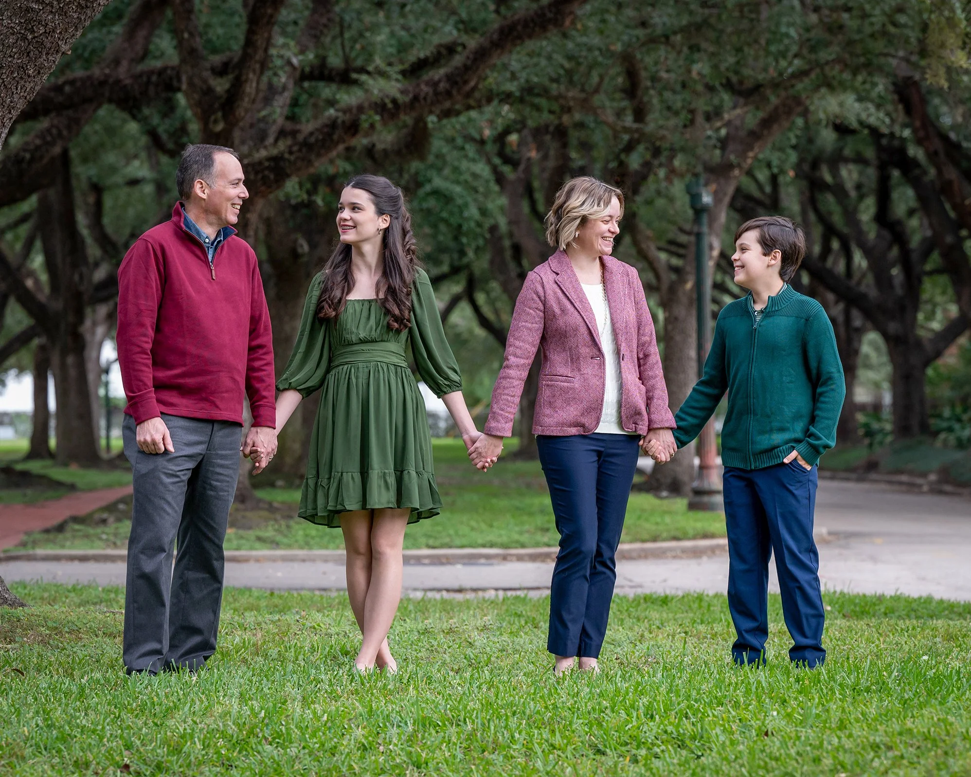 Family of four, two women and two men, holding hands and walking together outdoors in a park with large leafy trees.