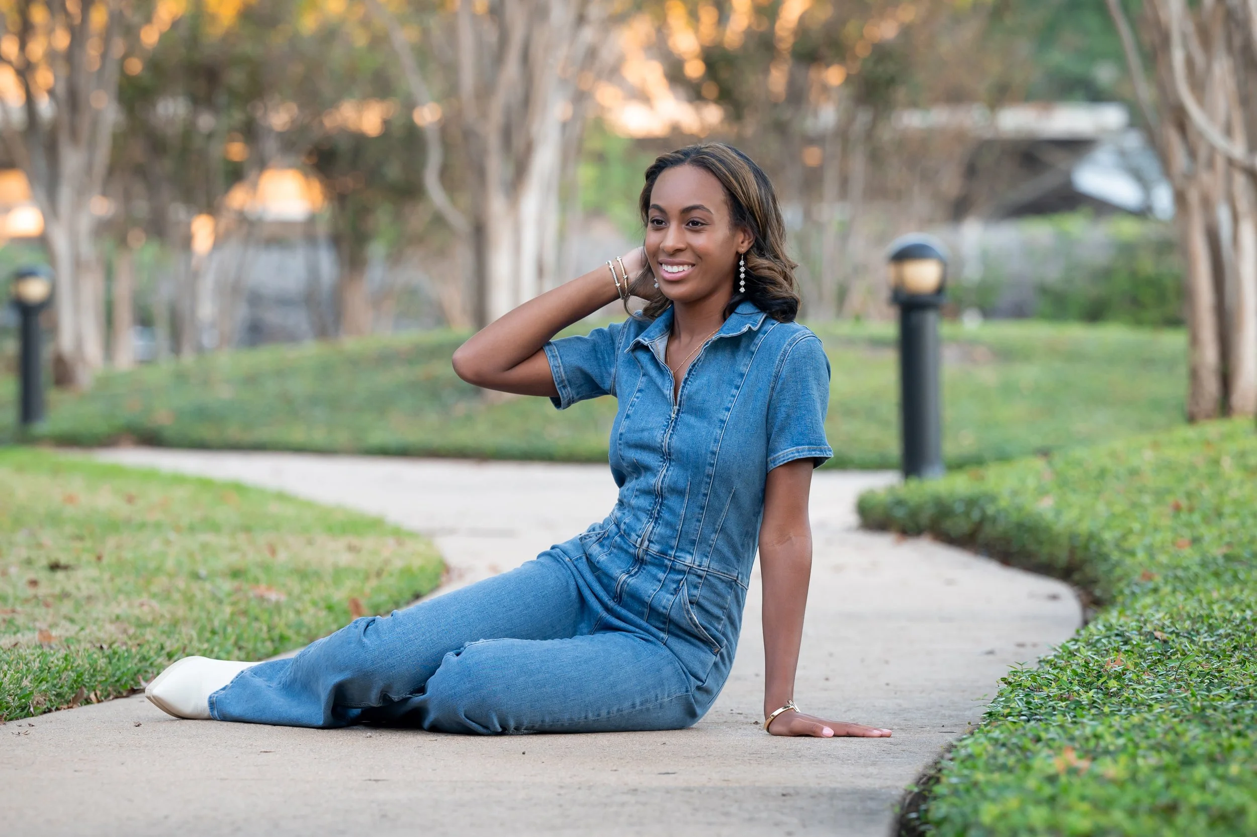 A woman in denim outfits poses on the sidewalk in a park setting with trees and grass, in a relaxed seated position with one hand behind her head and a smile.