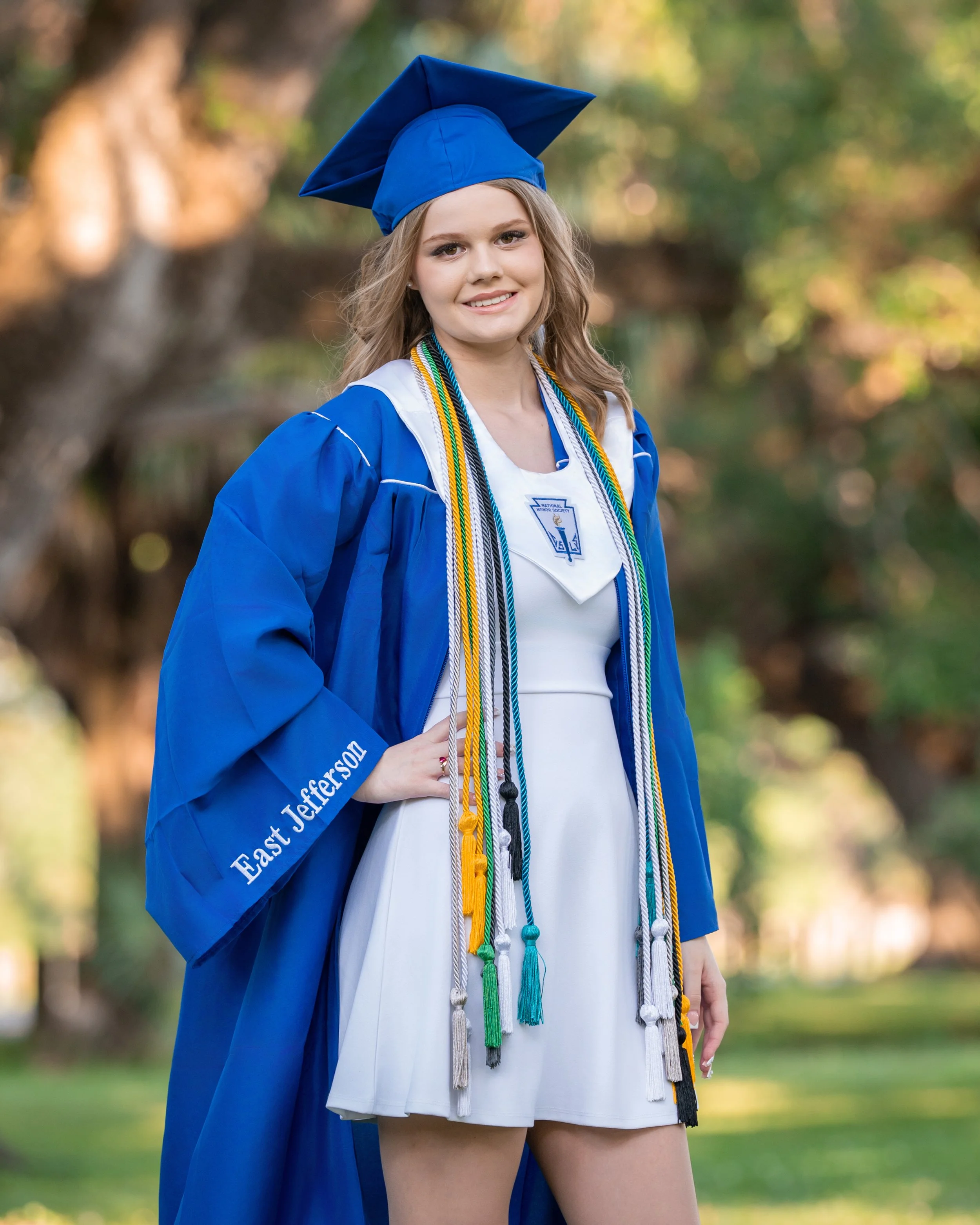 Young woman in a blue graduation gown and mortarboard cap standing outdoors, smiling, with colorful cords hanging around her neck, against a background of trees and grass.