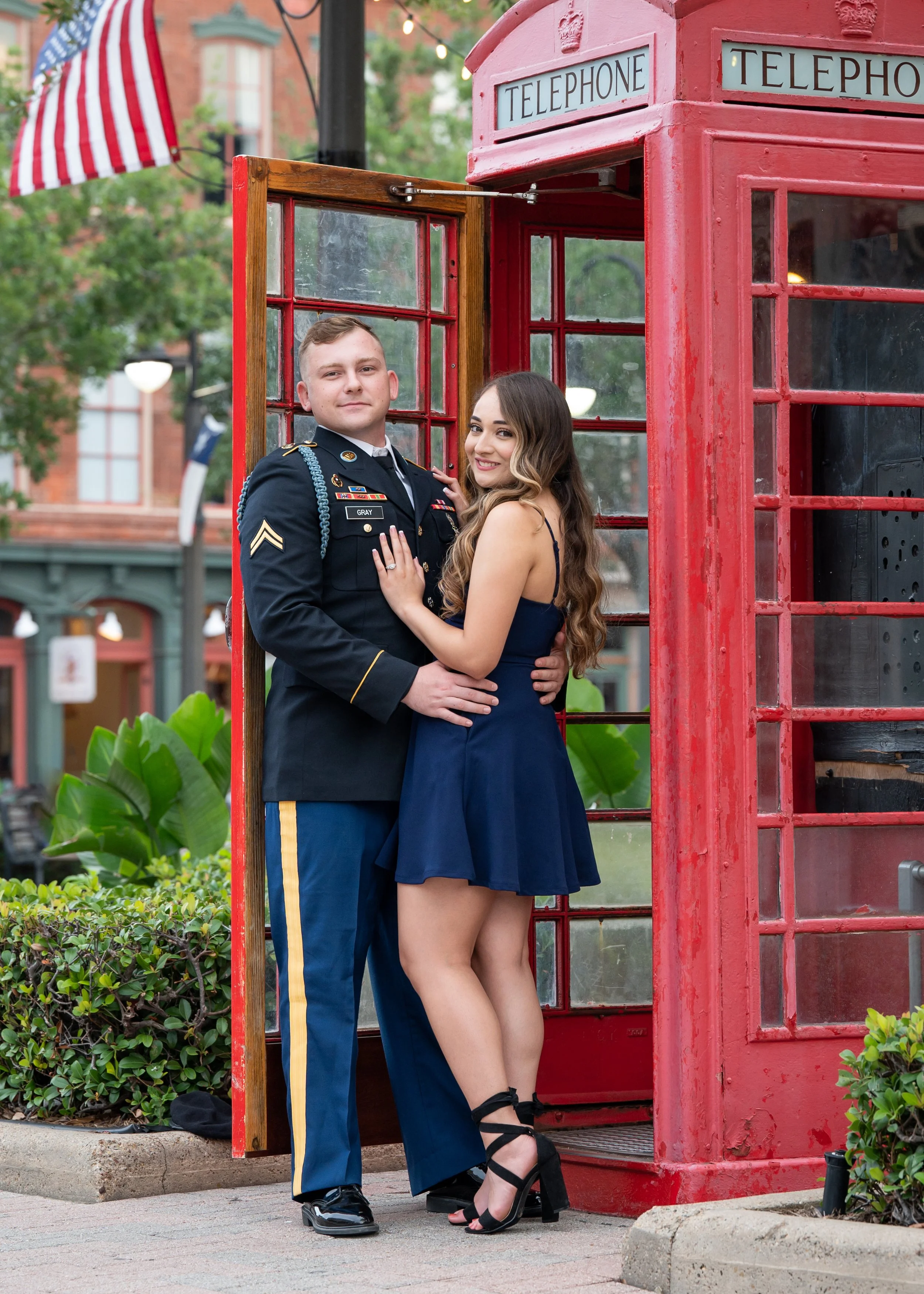 A young man in a military uniform and a young woman in a dark dress pose inside a red British telephone booth in an outdoor setting, with greenery and buildings in the background.