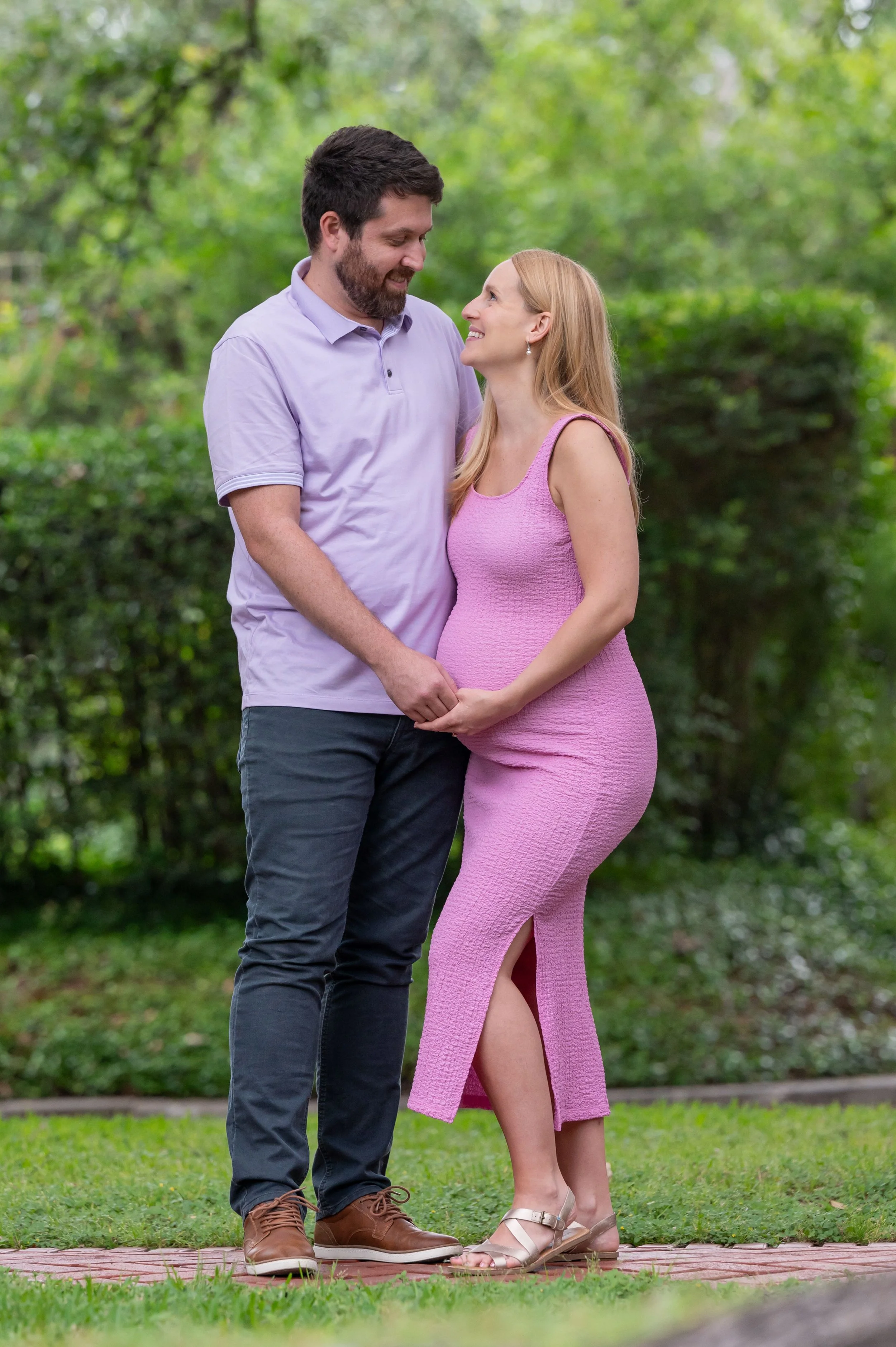 A pregnant couple, a man and a woman, standing close together outdoors on a brick path, holding hands, looking into each other's eyes with a green park background.