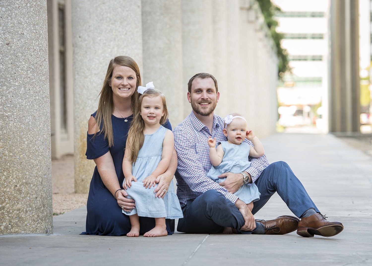 A happy family of four, including a woman, a man, and two young girls, posing outdoors on a sidewalk near large concrete pillars.