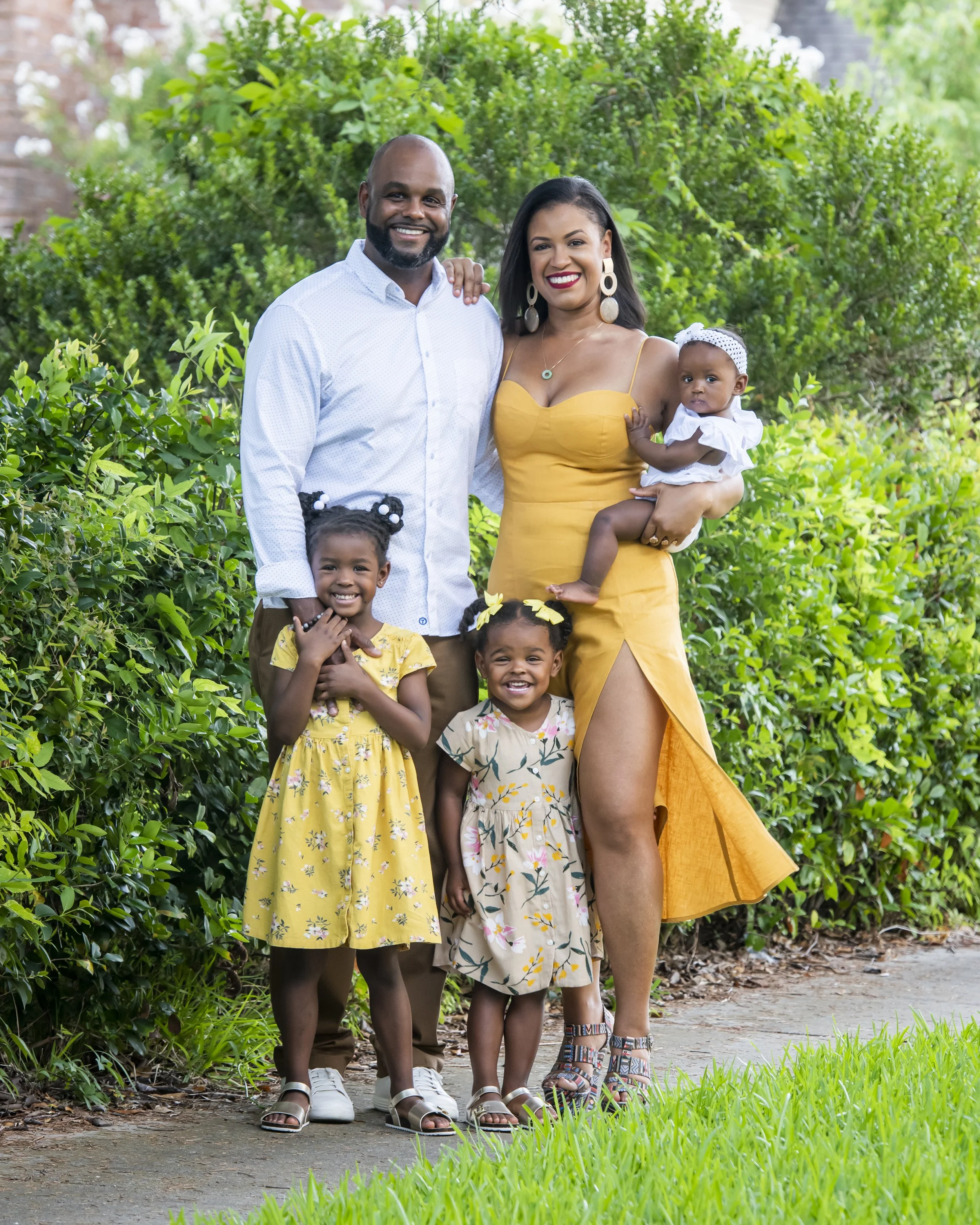 Family group photo of a man, woman, and three children standing outdoors on a nature trail, smiling. The man has a beard and is wearing a white shirt and brown pants. The woman holds a young girl and wears a yellow dress with large earrings. Two othe